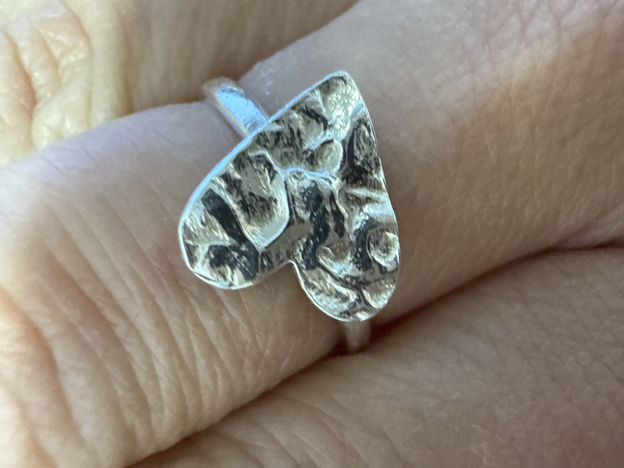 A close-up image depicts a silver ring with a heart-shaped setting, worn on a person's finger. The ring band is plain and thin, while the heart-shaped setting has a textured, hammered appearance. The finger appears to be wrinkled. There is no context to identify Tel-Aviv, Israel.