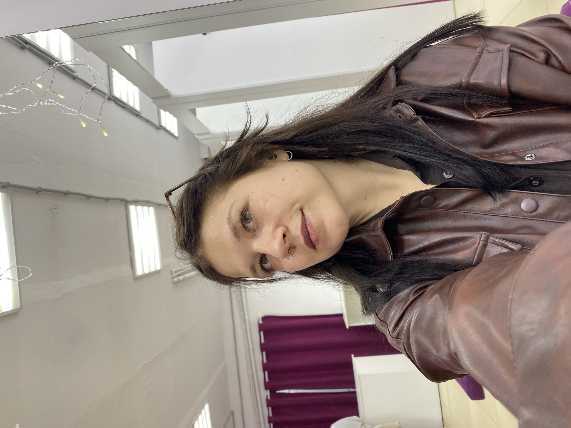 A woman with light skin and dark hair, wearing a brown leather jacket, stands indoors in a room with a window decorated with string lights. The room also includes purple curtains, white walls, and white furniture. This scene is likely in Chaikovsky, Russia.