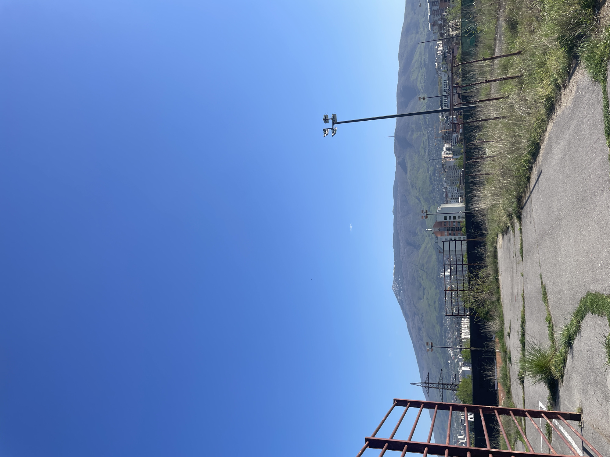 The image displays an outdoor landscape on a bright, clear day, set against the backdrop of a prominent mountain range. The scene suggests a neglected or disused area, likely in Sofia, Bulgaria, given the provided context.
In the foreground, a rusty, reddish-brown metal railing or gate structure is visible on the right. Much of the lower portion of the image is covered by a cracked asphalt or concrete surface, heavily infiltrated by tufts of grass and weeds growing through the fissures. This suggests the area was once paved, perhaps a road or court, but has fallen into disrepair.
The midground features several prominent man-made structures. A tall, dark-colored metal pole with multiple floodlight fixtures mounted at its top stands out, positioned to illuminate the ground below. Further back, several rusty metal posts, possibly remnants of an old fence, are partially obscured by tall, dry grass. A large metal electricity pylon or transmission tower is also visible, hinting at industrial or public utility infrastructure nearby. Buildings, characteristic of urban development, are clustered at the base of the mountains.
The background is dominated by a large, rolling mountain range, covered mostly in lush green vegetation, indicating fertile slopes. Some higher parts of the mountains show lighter patches, possibly bare rock or very sparse vegetation. The sky above is a vibrant, cloudless blue, save for one tiny, wispy white cloud near the center.
The overall impression is one of peaceful decay, where human structures are gradually being reclaimed by nature, all under a vast, open sky. There is no human activity visible in the static scene. The bright conditions suggest it is midday or early afternoon. No visible text is present.
