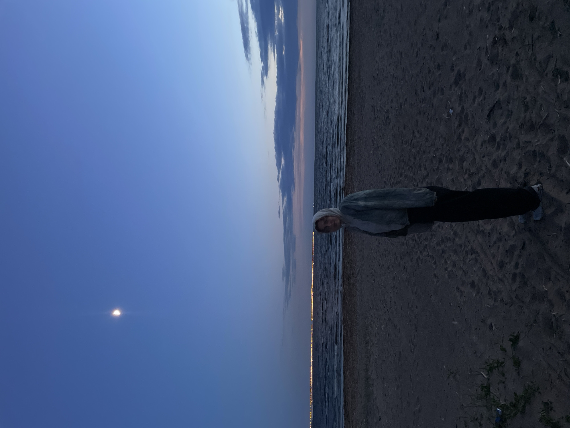 A vertical shot depicts a woman standing on a beach in Sestroretsk, Russia, at dusk. The sky transitions from dark blue on the left, where a crescent moon is visible, to lighter blues and a strip of faint pink on the horizon. Clouds are present along the horizon line. The body of water is to the left of the woman, with reflected city lights creating a thin golden line along the water's edge. The beach is dark sand. The woman is wearing dark pants and a light-colored jacket and head covering. She is facing the camera.