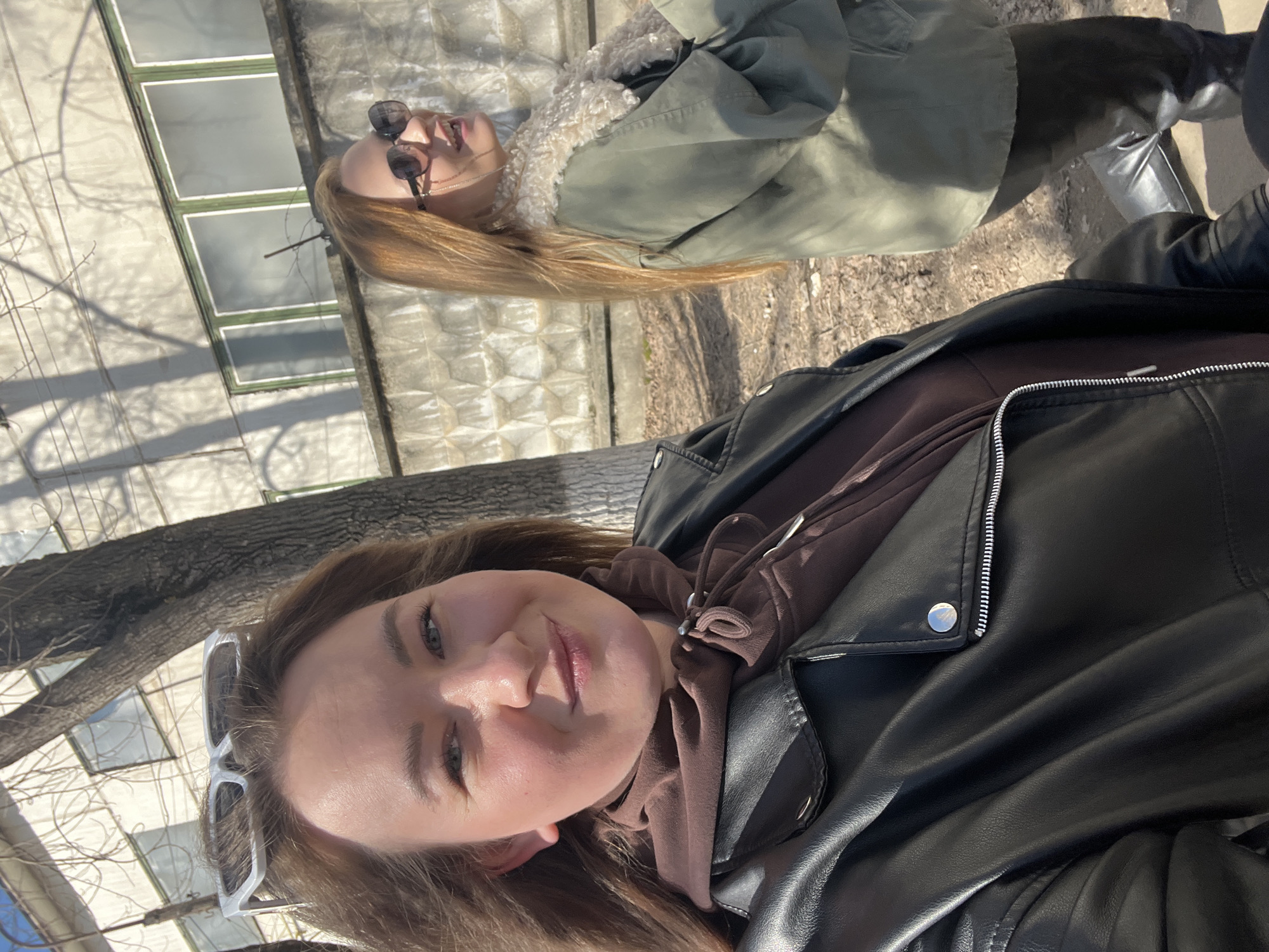 Two women are outdoors on a sunny day in what appears to be Chaikovsky, Russia, based on architectural styles. The woman in the foreground is wearing a black leather jacket over a brown hoodie and white-framed sunglasses. She is looking at the camera. The second woman is wearing an olive-green jacket with a furry collar, sunglasses, dark pants, and grey boots. They are both near a concrete building with windows. A bare tree is visible nearby.