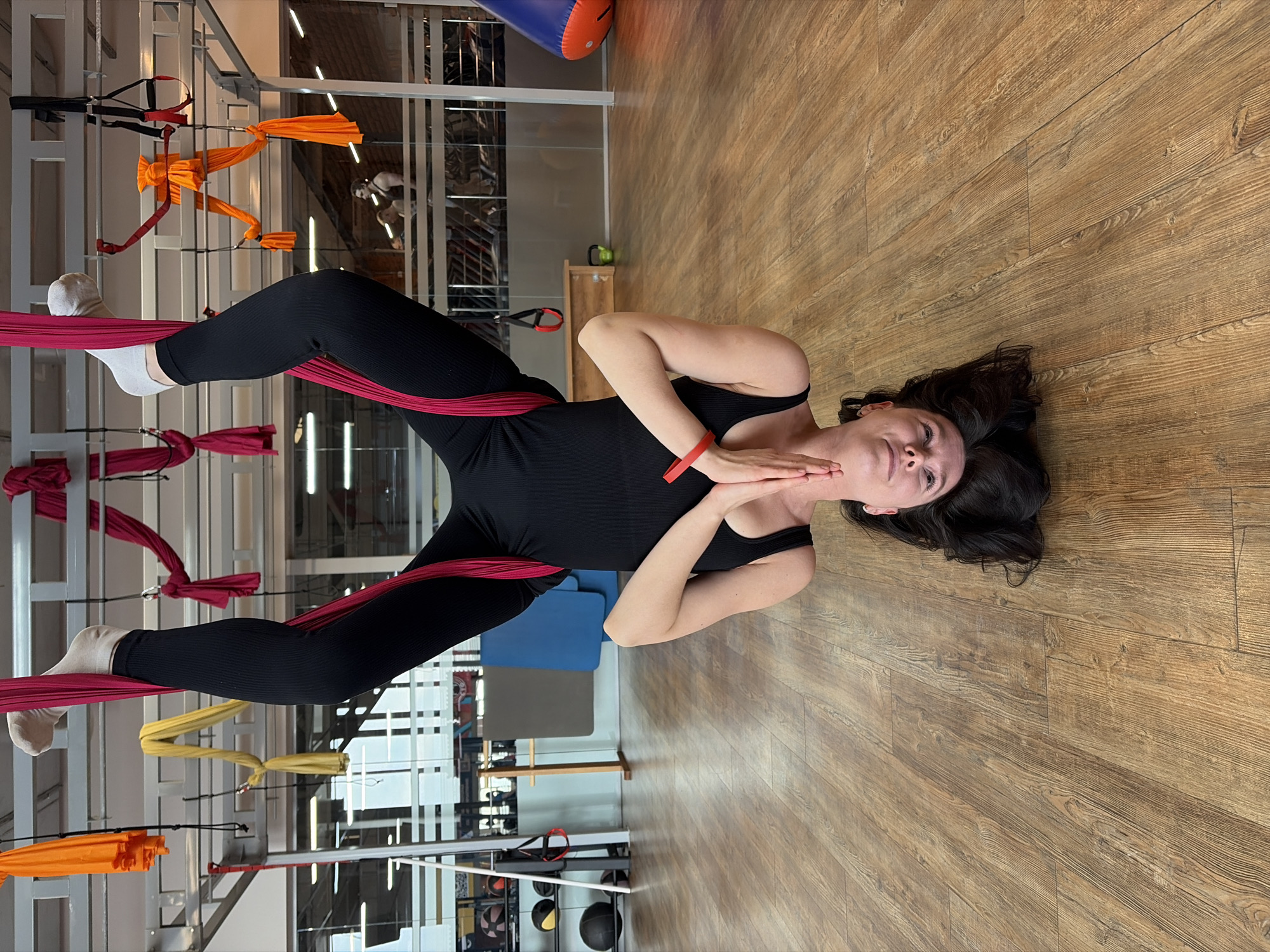 A woman in a black bodysuit and white socks is suspended in an aerial yoga hammock at a fitness center in Chaikovsky, Russia. The hammock is made of red fabric and is suspended from a metal frame. The woman is lying on her back with her hands clasped near her neck. The floor is made of brown wood. Various gym equipment and apparatus are visible in the background.