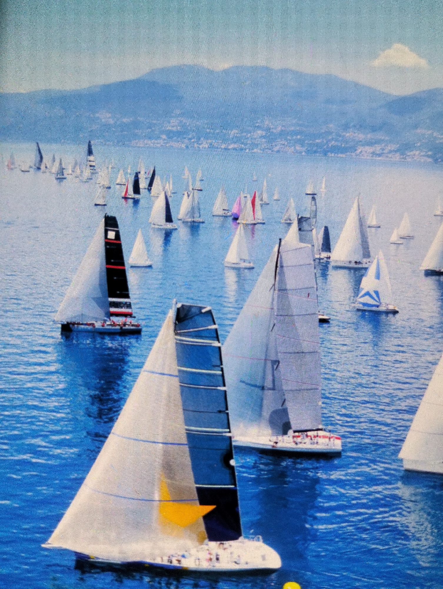 This is a painting depicting a sailing regatta. A large fleet of sailboats with white and dark-colored sails navigate on a deep blue, choppy body of water. In the foreground, two sailboats are prominent. The one on the left has a partially visible sail with blue lines and a yellow triangular accent. The sailboat on the right has a distinct black and red striped sail. In the background, numerous other sailboats are scattered across the water, appearing smaller in scale. A hazy, light blue sky stretches above, with a faint white cloud visible. Distant, soft blue mountains with a hint of urban development line the horizon. The image does not provide any information about the city of Payagaboh, Indonesia.