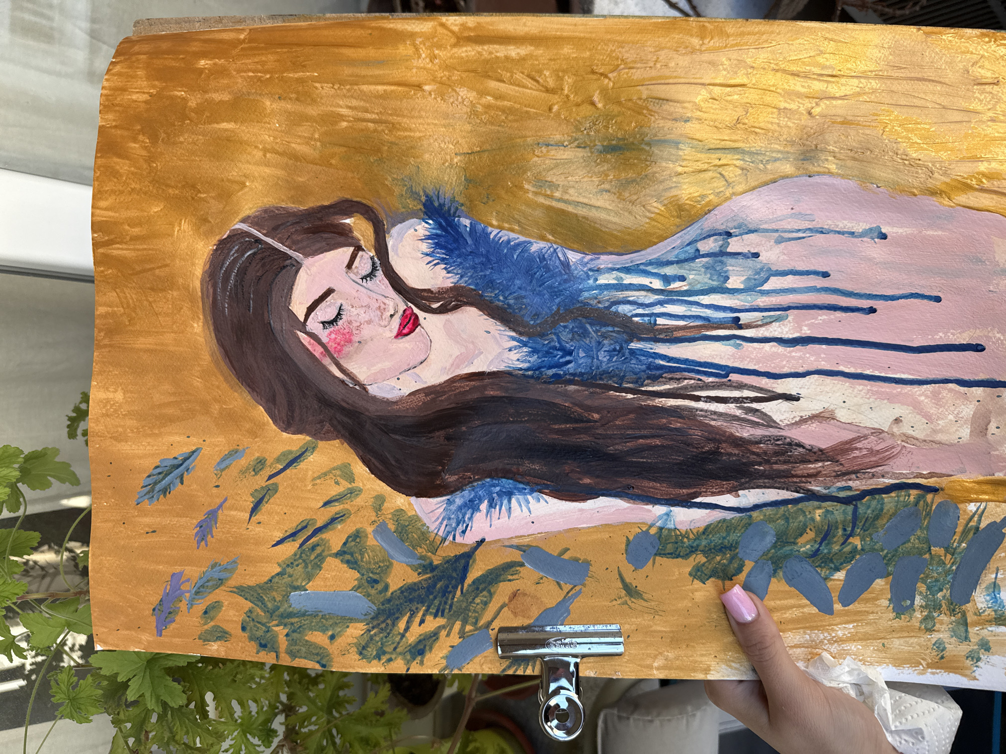 The image shows a painting of a woman lying down. She has long brown hair and fair skin. She is wearing a light pink dress with blue feather-like embellishments. The background is gold. The painting is being held up by a hand with pink nail polish. There is a plant and a clamp visible in the foreground. The location of the image is unknown.
