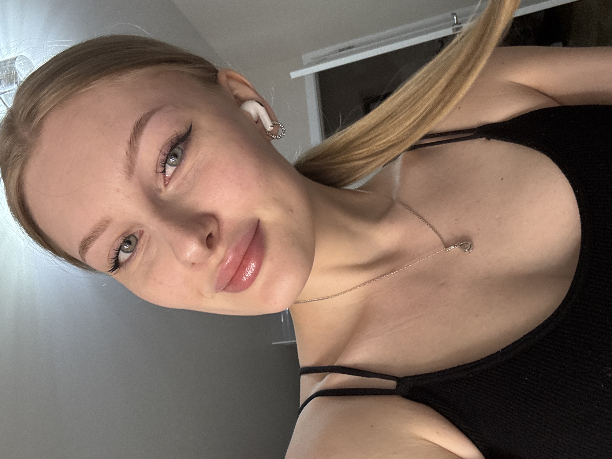 A young woman is seen in a selfie-style photograph. She has fair skin, blonde hair pulled back in a ponytail, and light-colored eyes. She is wearing a black tank top with thin straps and a delicate silver necklace. An earbud is visible in her right ear, along with a small hoop earring. The background is a plain gray wall and a partially visible white door or frame. The exact location within Perm, Russia cannot be determined based on the provided image.