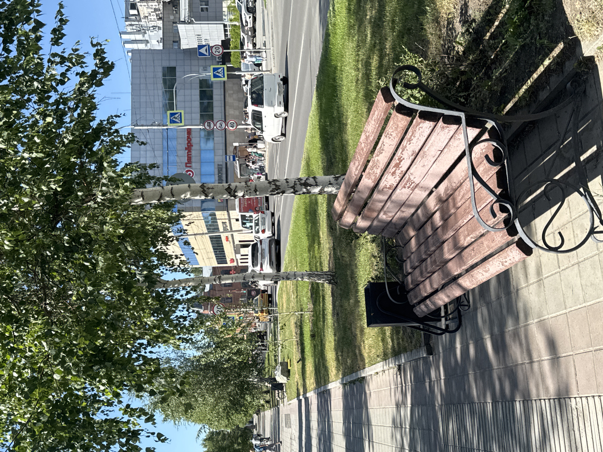 A sunny day scene unfolds along a paved pedestrian walkway bordering a grassy median. A weathered wooden bench with a black metal frame sits in the foreground, casting shadows on the walkway. Beyond the median, a multi-lane road carries vehicular traffic, including cars, a white van, and a bus. Buildings line the far side of the road, one with a prominent sign that reads "Пятёрочка" (Pyaterochka), a Russian supermarket chain. Several pedestrians are visible, some waiting at a bus stop, others walking along the sidewalk. Traffic signs, including speed limit indicators (20 and 40), and pedestrian crossing signs are present. The environment suggests an urban setting, specifically identified as Barnaul, Russia, based on the presence of the "Пятёрочка" store. The overall atmosphere is of a typical day in a Russian city.