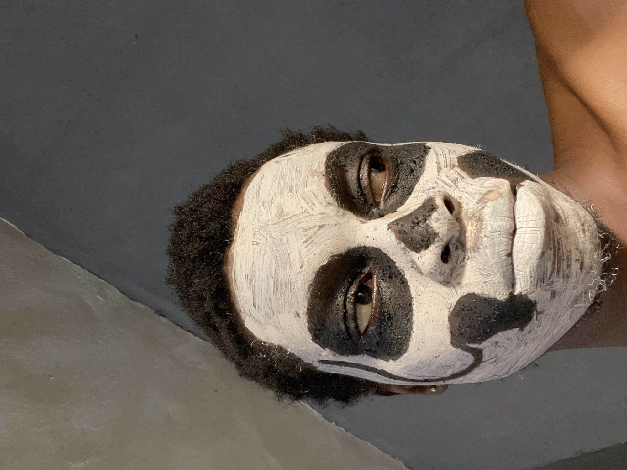 An eye-level, close-up shot captures a person with intricate face paint, likely posing for a photograph. The subject's head is tilted to their right, and their eyes are open and looking directly ahead with a neutral expression. Their face is covered in thick, textured white and black paint, creating a design that resembles a skull or skeleton. White paint covers the majority of the face, applied with visible brush strokes, while black paint forms large, dark circles around the eyes, extends down the bridge of the nose, and creates a stylized design on the cheek and around the mouth area.
The person has short, dark, curly hair framing their face. Their skin tone on the visible neck and shoulder area is warm brown. The background is simple and uncluttered, featuring a plain, dark gray surface behind the head and a lighter gray or beige surface in the bottom left, possibly indicating a wall and floor or another wall in an indoor setting. The lighting is soft and even, highlighting the texture of the face paint. There is no visible text.