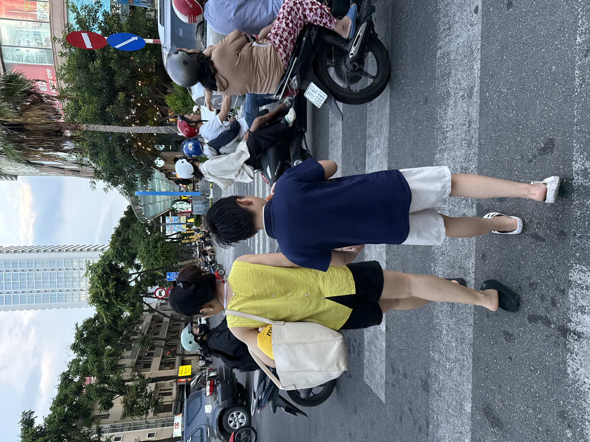 A woman and a young boy are walking across a crosswalk in Nha Trang Ward, Vietnam. The woman is wearing a yellow sleeveless top and black shorts, carrying a large beige tote bag. The boy is wearing a navy blue t-shirt and white shorts. They are walking away from the camera, towards the right side of the image. Several motorbikes are visible in the background, some with riders. The environment appears to be a city street with buildings and trees lining the road. There are traffic signs and streetlights present. The sky is overcast, suggesting it is either early morning or late afternoon.