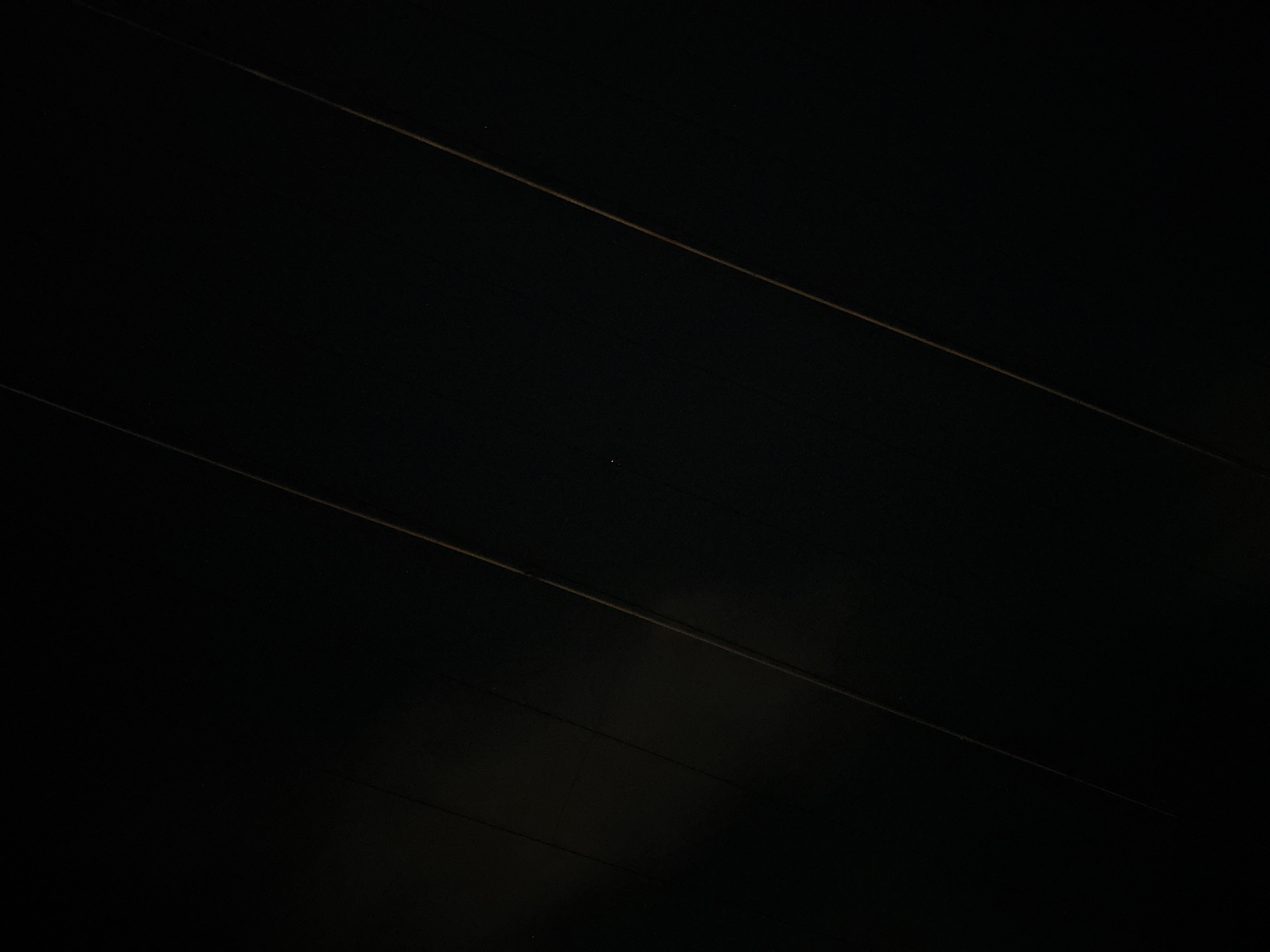 The image is a close-up, low-light photograph of what appears to be a dark, textured surface, possibly a ceiling or wall paneling, with subtle diagonal lines of light or reflection running across it. There is a small, faint point of light visible near the center of the image, which could be a distant star or a light source. The overall impression is one of darkness and abstract geometry, with no discernible elements that would indicate a specific location in Nha Trang, Vietnam, or any people, objects, or actions. The image is purely descriptive of a visual texture and faint celestial or artificial light.