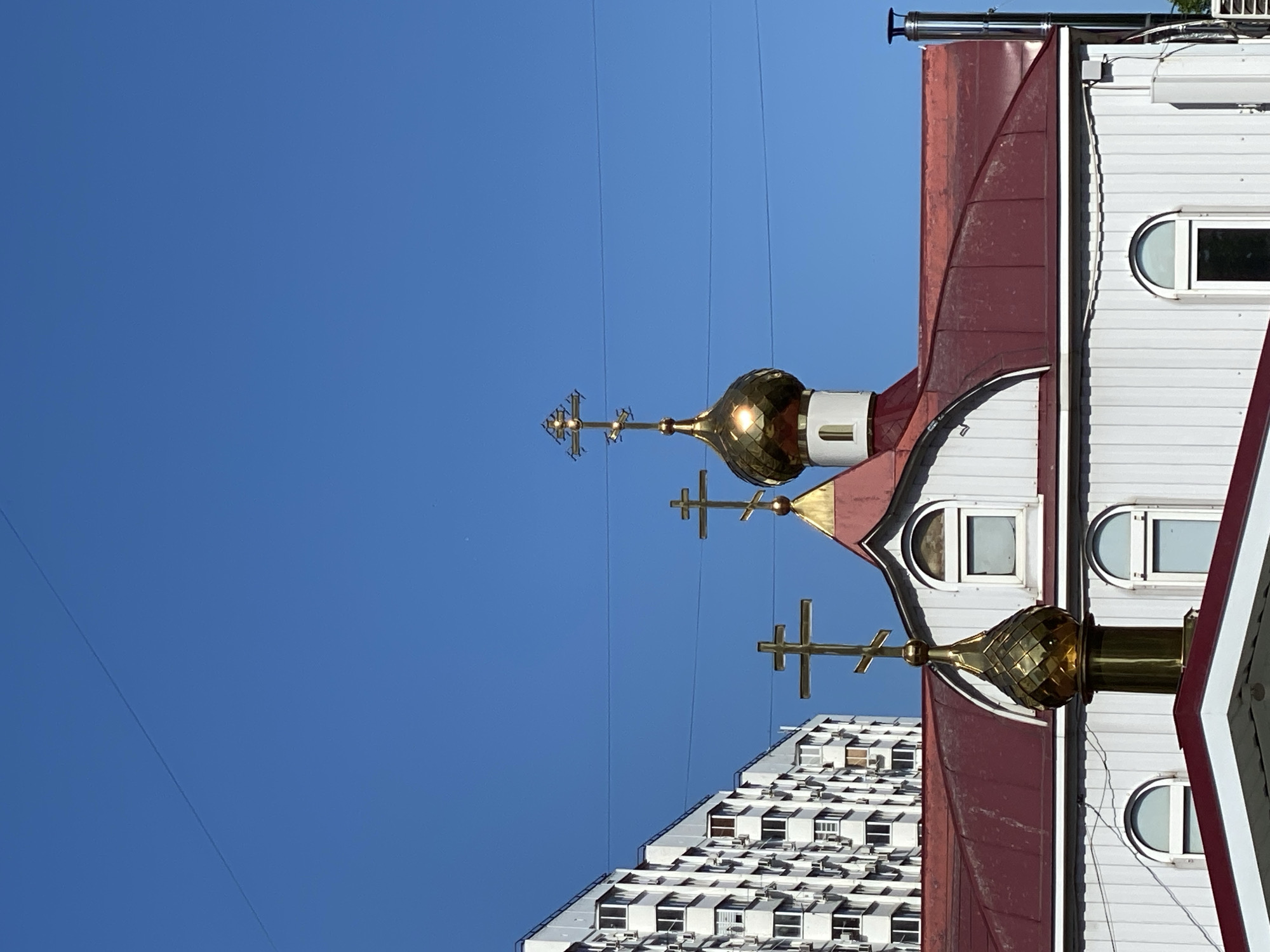 The image shows the upper section of a building, likely a church, with two golden domes and crosses against a clear blue sky. Red roofing and white siding are visible on the building's exterior. Adjacent to the church is a large white residential building with numerous windows, suggesting an urban environment. Power lines can be seen traversing the sky.