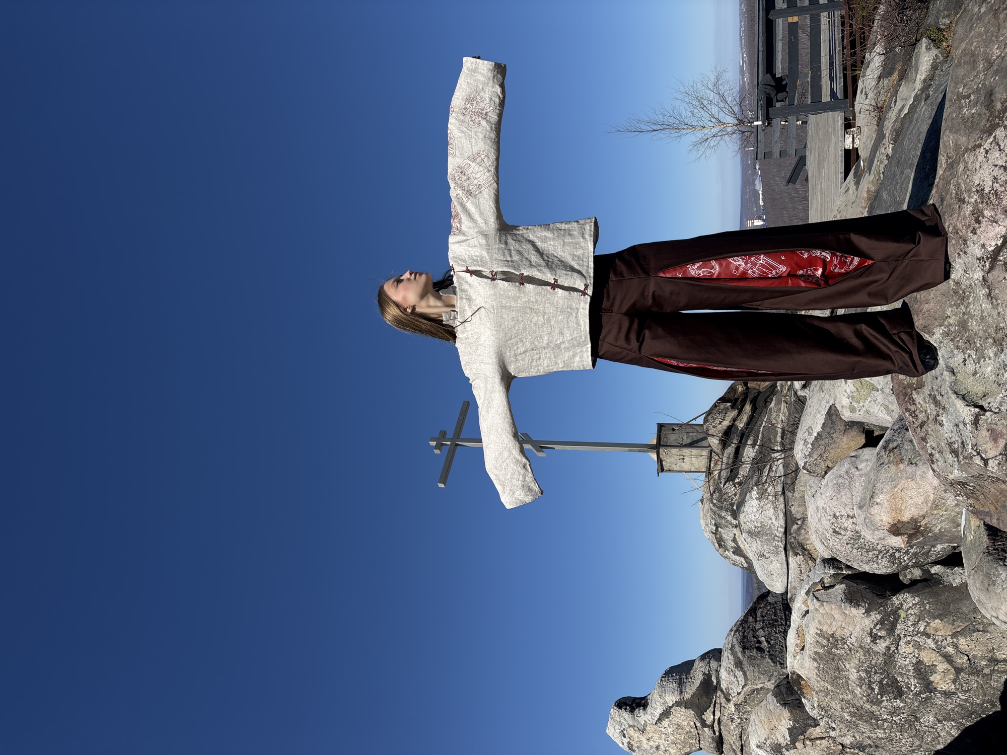 A young woman stands on a rocky summit in Gubakha, Russia, facing the sky with arms outstretched in a cruciform posture. A metal cross is visible nearby. She is wearing brown trousers with red patterned insets and a light-colored, patterned jacket. The sky is a clear, deep blue. In the background, a railing and a glimpse of a distant landscape are visible.