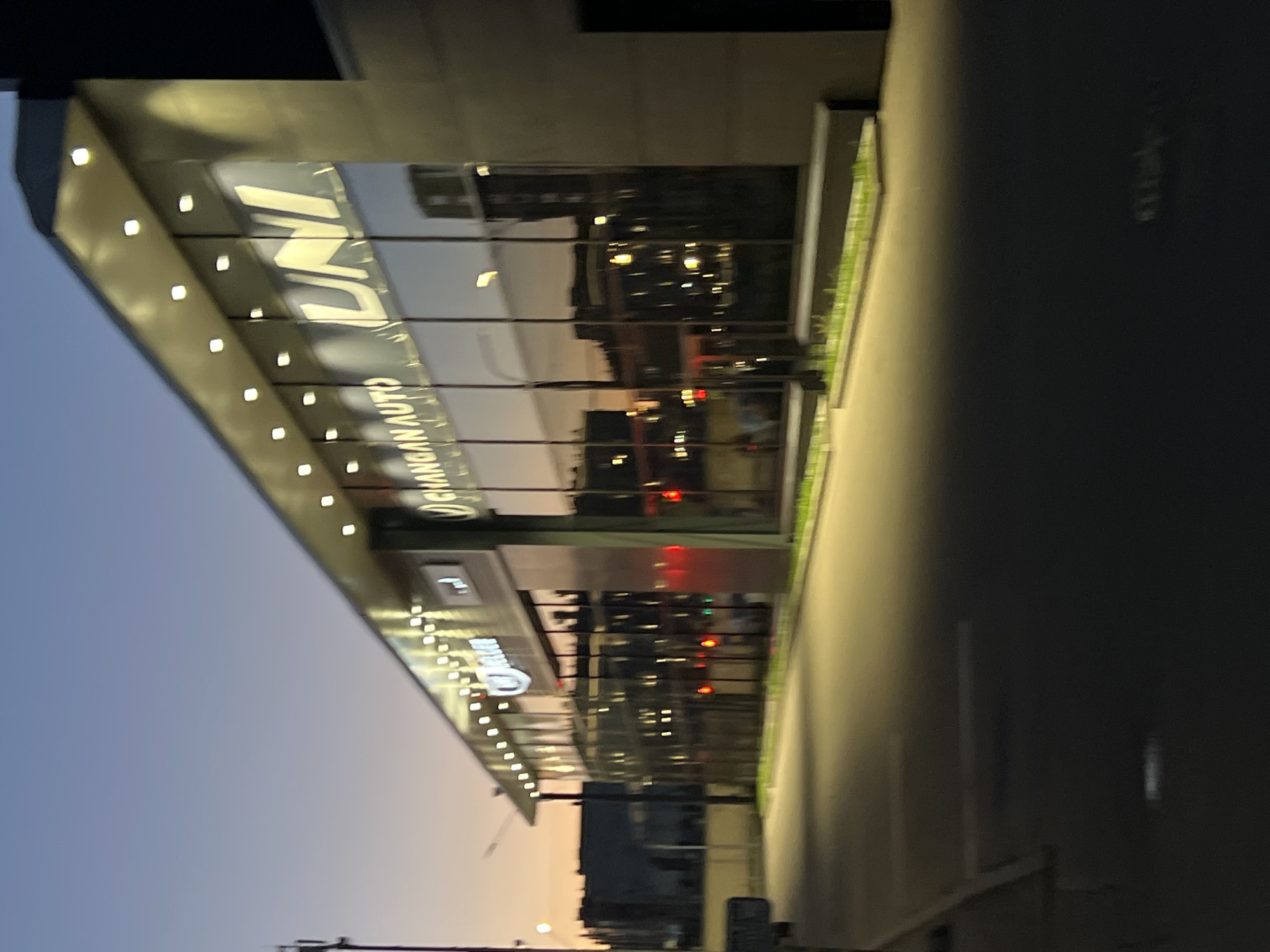 This image depicts the exterior of a modern commercial building at dusk in the city of Unknown, Unknown. The structure features a prominent glass facade illuminated by numerous square-shaped, recessed lights along its roofline. Large, white, illuminated signage on the glass displays "CHANGAN AUTO" on the left side and "UNI" in stylized capital letters on the right.
The glass facade reflects the surrounding environment, including a fading twilight sky with a gradient from blue to orange-pink, several distant urban buildings, and multiple scattered artificial lights, some appearing as red circular traffic signals.
In the foreground, an asphalt surface, likely a parking lot or road, is partially illuminated by the building's lights, casting a yellowish hue. A curb runs along the base of the building, accompanied by a linear planter containing green vegetation. The background reveals additional dark, silhouetted buildings against the twilight sky, and a dark utility pole is visible on the far left. No individuals or vehicles are present in the visible area. The overall image exhibits a slight softness or out-of-focus quality.