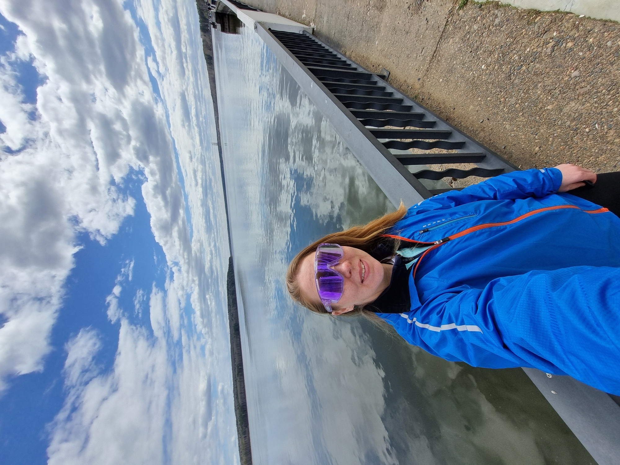 A woman is pictured in a selfie against a backdrop of the Kama River in Chaikovsky, Russia. She wears a blue jacket with orange trim and reflective, likely purple, sunglasses. Her red hair is visible. The river, reflecting the sky, stretches out behind her, with trees lining the far bank. To her right, a metal staircase and a concrete walkway are visible. The sky is partly cloudy with blue patches.