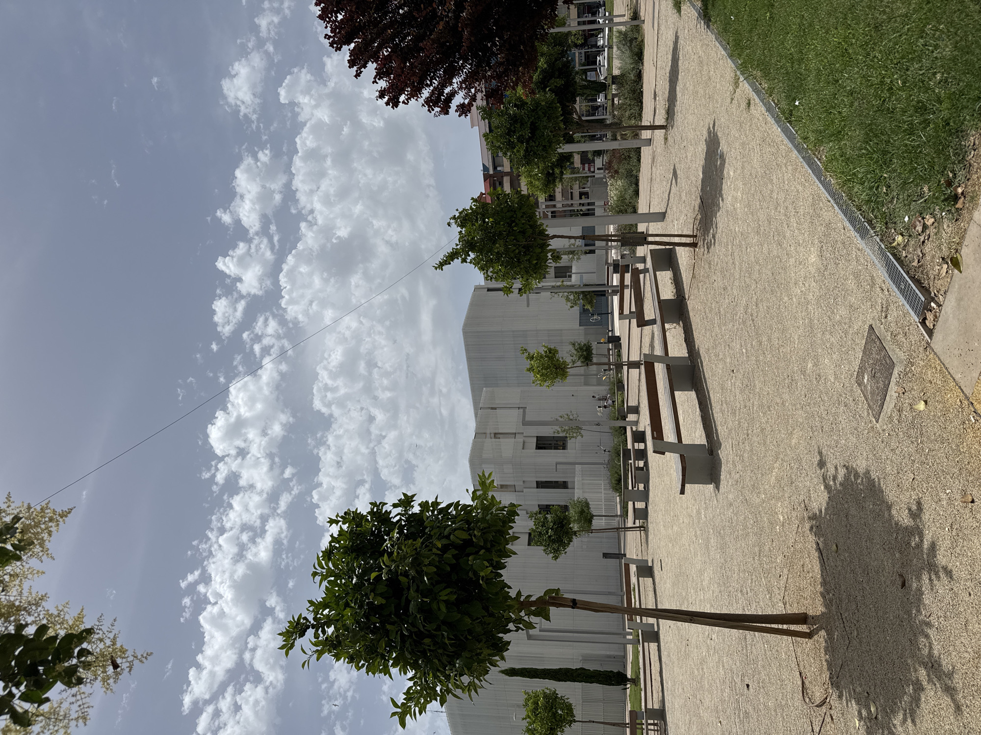 The image depicts a modern urban park with a contemporary building in the background. The park features a gravel path lined with trees and benches, creating a serene ambiance. A lush green lawn borders the path, contrasting with the neutral tones of the urban landscape. The sky is partly cloudy, suggesting a pleasant day, likely late morning or early afternoon. The architecture of the building is geometric and minimalist, with vertical lines and a pale facade. The trees are neatly planted, some with full green canopies, others in the early stages of growth. Shadows cast by the trees and benches indicate the sun is present, illuminating the scene. There is no visible text in the image. The setting appears to be a well-maintained public space designed for relaxation and leisure, possibly in a city in Spain like Gandia. The overall mood is tranquil and orderly.