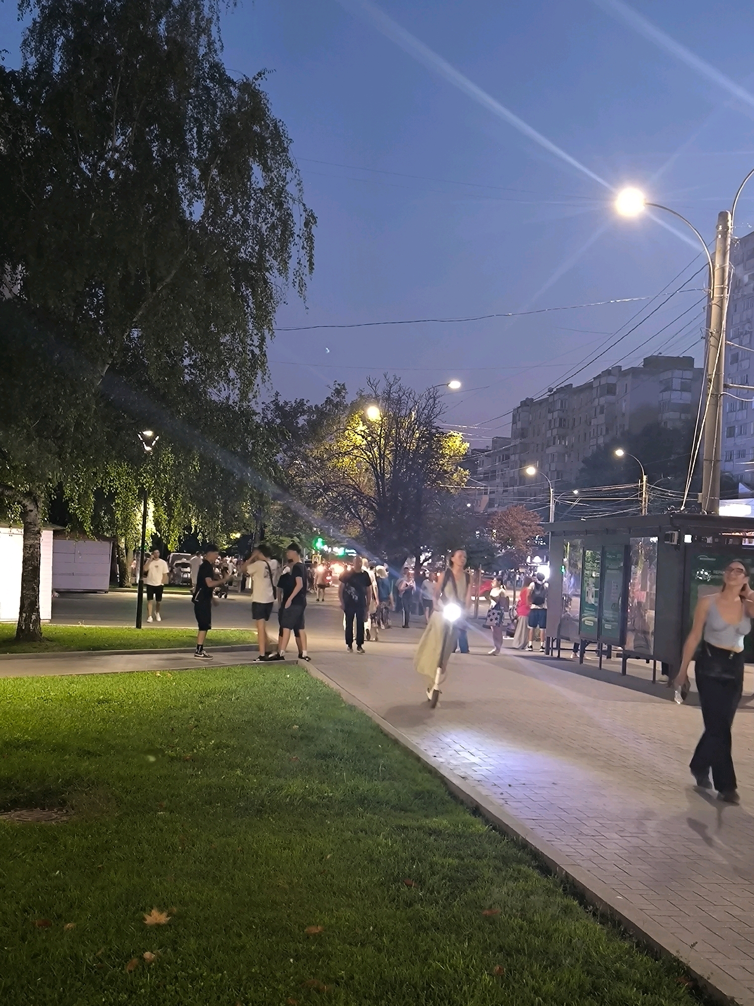 The image depicts a pedestrian area in Chișinău, Moldova, at dusk. A paved walkway is lined with green grass on the left. Several streetlights are visible, with one in the foreground casting a bright light on the walkway. A young woman is riding a scooter on the right side of the walkway, illuminated by a streetlight. On the left, a group of young men are standing and conversing near a tree. In the background, apartment buildings and a bus stop are visible. The sky is a deep blue, with the hint of a crescent moon. Several vehicles and other pedestrians are present further down the walkway.