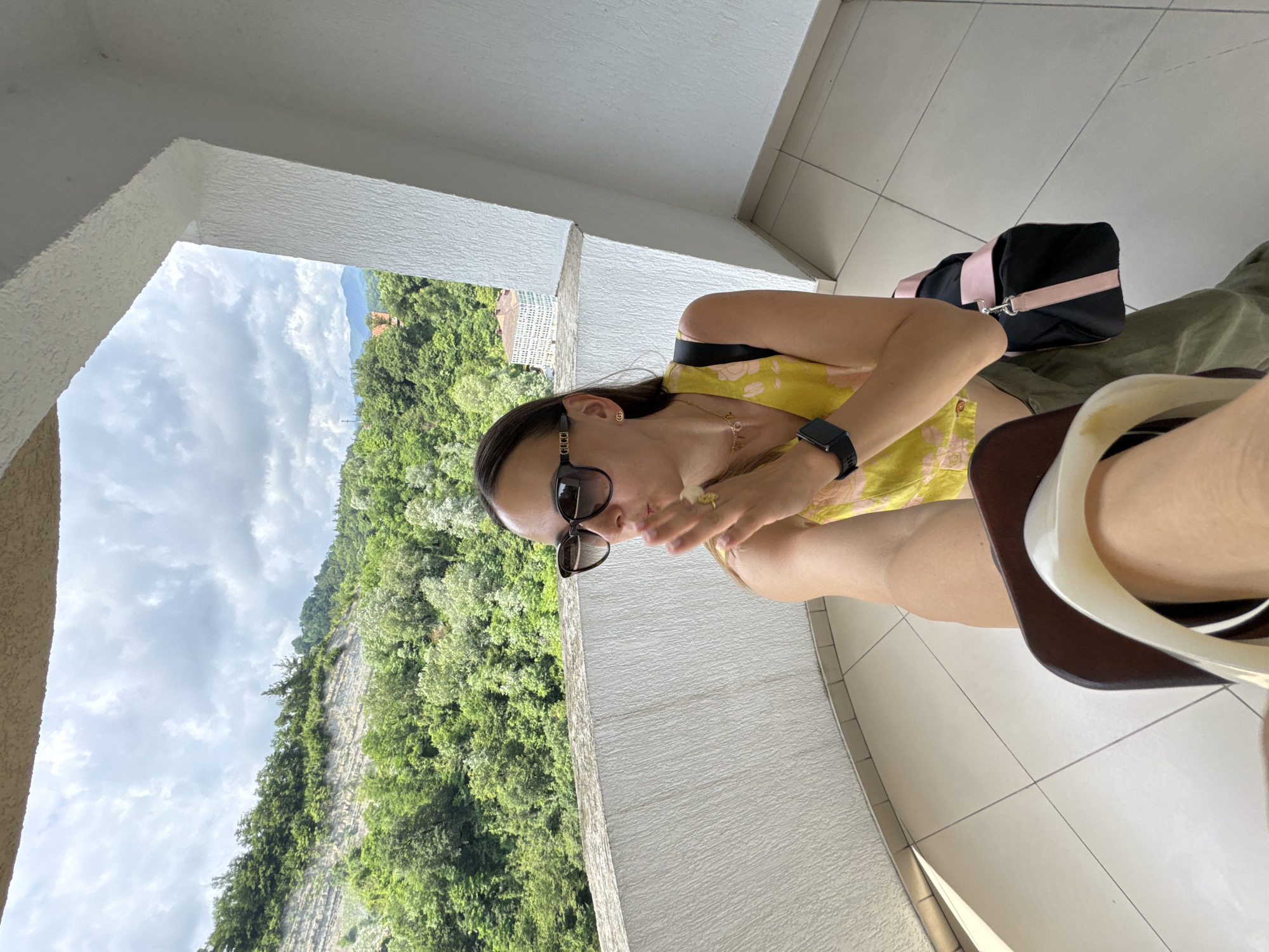 This is a portrait of a woman sitting on a balcony with a lush green mountainous landscape visible in the background. The woman is wearing a yellow floral top, sunglasses, and a watch on her left wrist. She has a ring on her right hand and is holding her hand up to her face. A black and pink duffel bag is placed behind her. The setting appears to be outdoors on a sunny day with some clouds in the sky. The architecture suggests a resort or hotel in Sochi, Russia, with tiled flooring and white textured walls. The woman's pose and the overall lighting suggest a relaxed moment, possibly during a vacation.