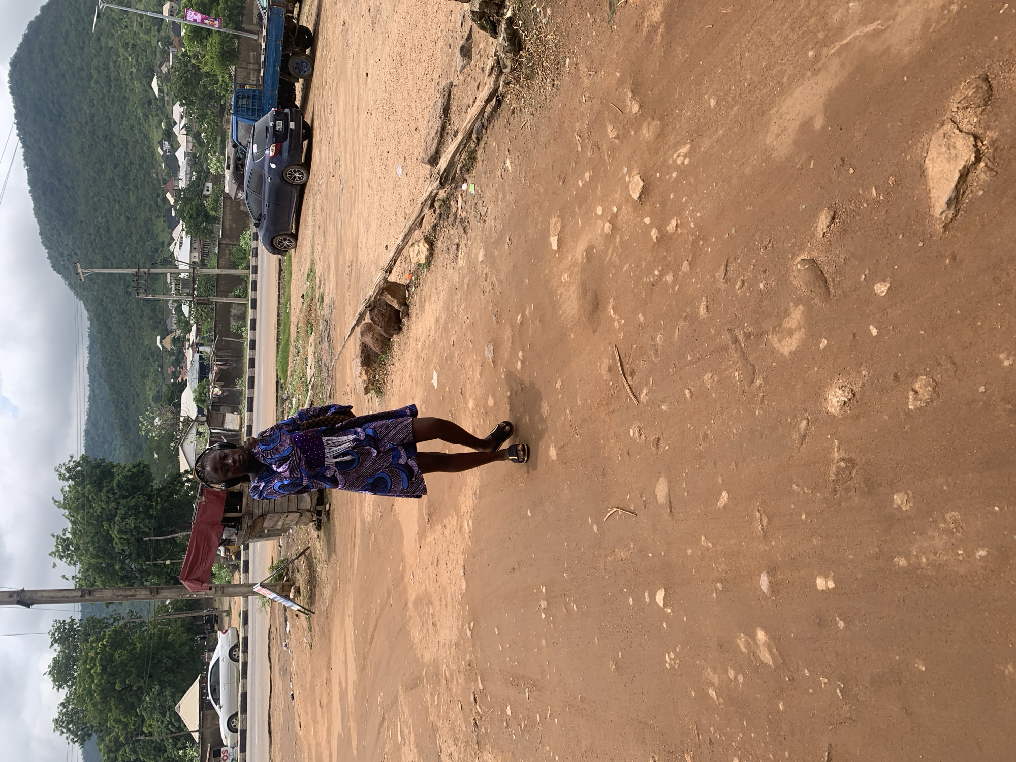The image depicts an outdoor scene in Adankolo, Nigeria. A young woman wearing a purple and blue patterned dress and black sandals stands in the foreground on a dirt road. She is looking towards the camera and holding a brown bag. She is also wearing headphones. In the background, there is a utility pole, a small market stall with a red awning, a white car, and other vehicles including a blue flatbed truck. Beyond the road, there are houses nestled on a green, mountainous landscape under a cloudy sky. The ground is uneven and composed of reddish-brown dirt with scattered rocks and debris.