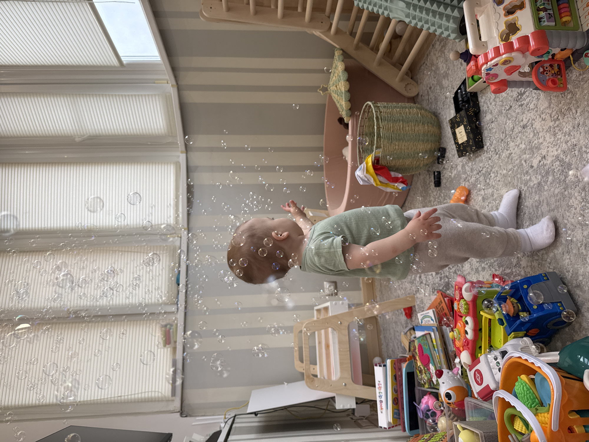 The image depicts a child indoors playing with bubbles. The child is wearing a green shirt and gray pants with white socks. They are surrounded by various toys including a wooden climbing structure, a toy basket, a blue and yellow toy truck, books, and other play items. The background includes a window with white blinds, a striped wallpaper, and a gray carpet. The scene is filled with bubbles of various sizes. There is no evidence of the image location being Moscow, Russia.