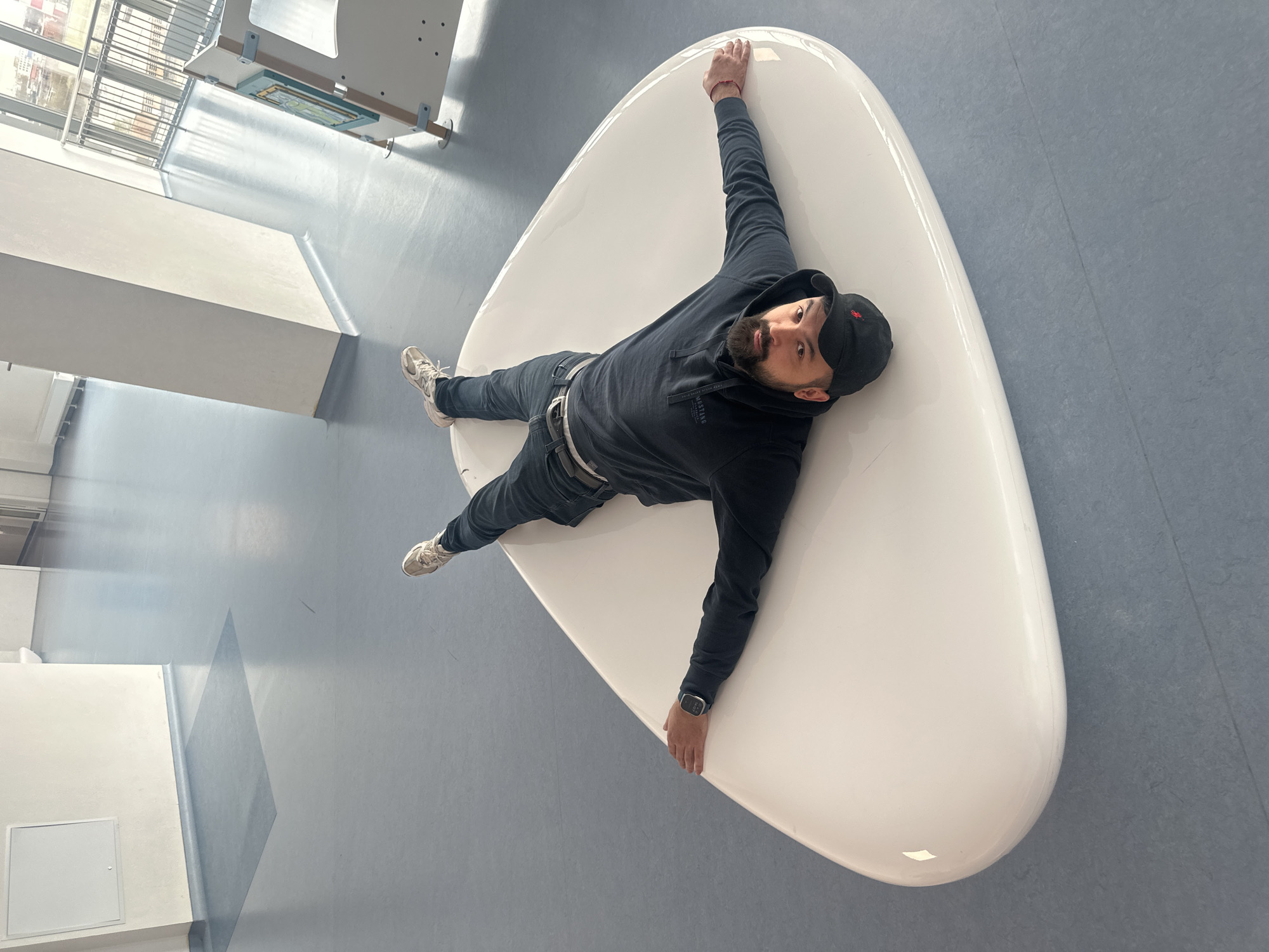 An overhead shot, slightly tilted, captures a man lying supine on a large, smooth, kidney-bean-shaped white lounge in a bright, modern indoor space. The man, with a beard and wearing a black hoodie (with "MUSTANG" partially visible), dark jeans, white sneakers, and a black baseball cap with a small red logo, looks directly at the camera with a neutral expression. His arms and legs are outstretched, with a smartwatch visible on his left wrist and a red bracelet on his right.
The setting features a light blue floor with subtle darker blue square patterns and reflections from a nearby window. White walls and pillars, along with a large window showing horizontal bars and distant structures, suggest a contemporary public building, possibly a university, office, or waiting area. A white access panel is visible on the floor or a low wall. The space is well-lit, indicating daytime. This scene is set in Khimki, Russia.