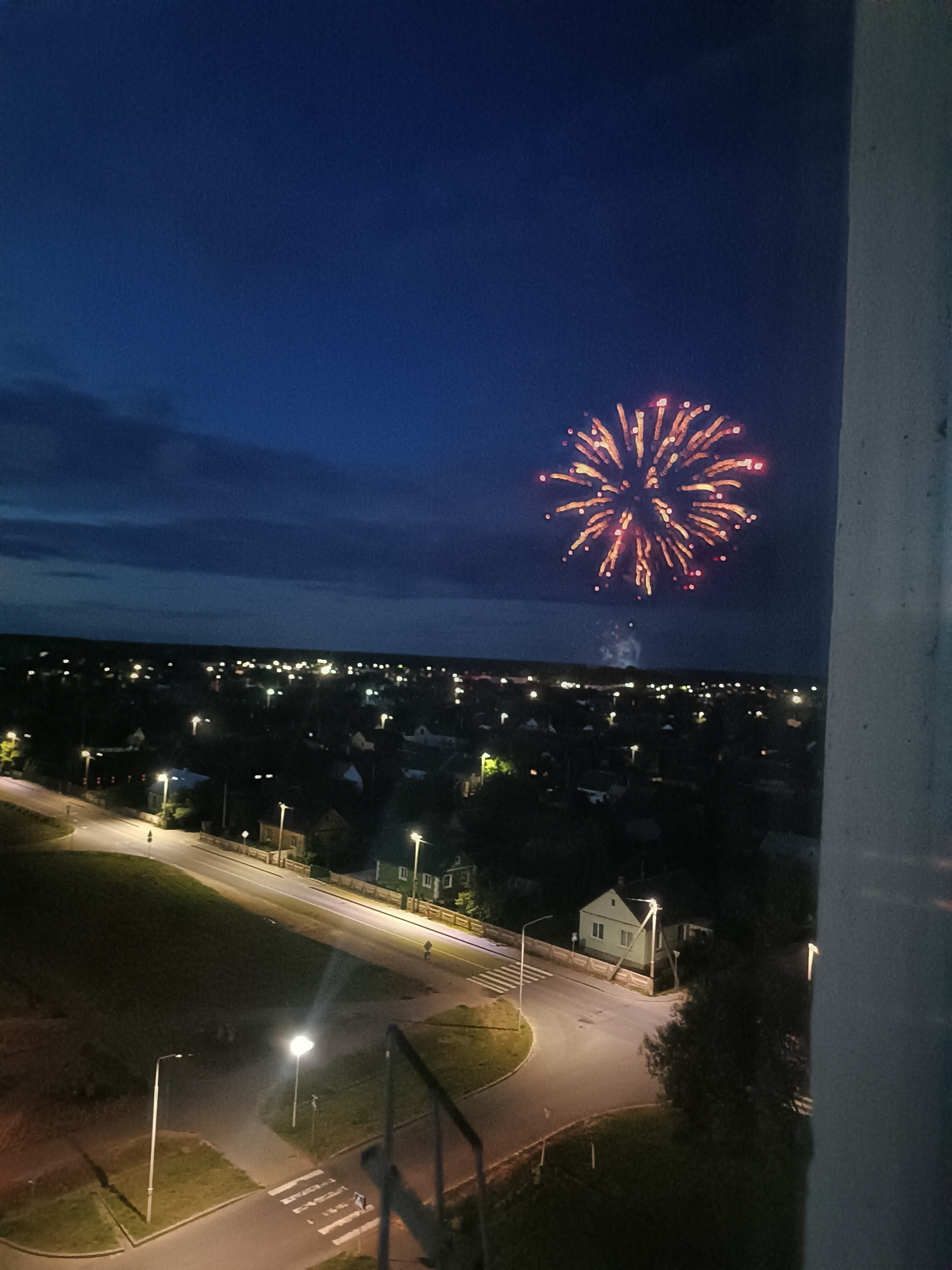 The image depicts a nighttime scene overlooking a residential area. A large, bright fireworks display is visible in the dark blue sky, with streaks of orange and red emanating from a central point. Below the fireworks, a town or village is illuminated by scattered streetlights and the lights of houses. The houses appear to be single-family dwellings with pitched roofs. Roads, some with streetlights, wind through the residential area, with green spaces and trees interspersed. A white pillar or wall is visible in the foreground on the right side of the image, suggesting the photograph was taken from a building. There are no people or animals visible in the frame. The overall impression is of a festive or celebratory event taking place in a quiet, suburban setting at night. The location is identified as Skidzieĺ, Belarus.