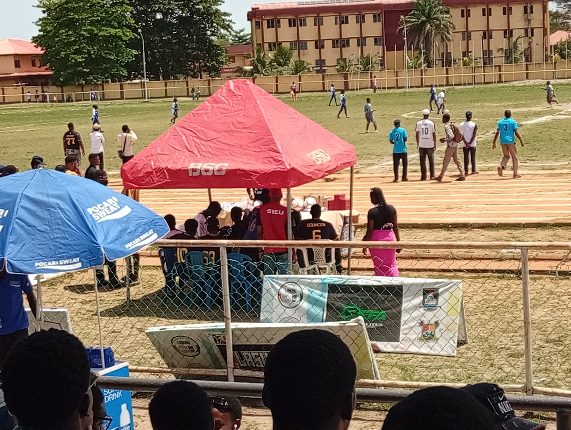 Here is a detailed description of the image:
The image shows a scene at what appears to be a sports event, likely a football (soccer) match, taking place in Lagos, Nigeria. The environment includes a grassy field where a match is in progress. Spectators are present, viewing the game from behind a metal fence.
Visible elements include:
* **People:** Players on the field, some wearing numbered jerseys (such as 77, 10, 3), and spectators. Individuals are seen wearing t-shirts with the word "SIEU" on them.
* **Objects:** Two tents. One is a red tent with writing on its side, and the other is a blue umbrella with "POCARI SWEAT" branding, used to provide shade. Balls are seen inside the red tent. Banners with logos or advertisements.
* **Actions:** Football (soccer) game is going on. People appear to be watching and interacting.
* **Environment:** The background includes a multi-story building and some trees. The event appears to be happening outdoors on a sunny day.
Other notable features:
* A cooler with "SOFT DRINK" visible on its side is present, suggesting refreshments are being sold.
* Several balls, presumably footballs, are visible near the tent.
