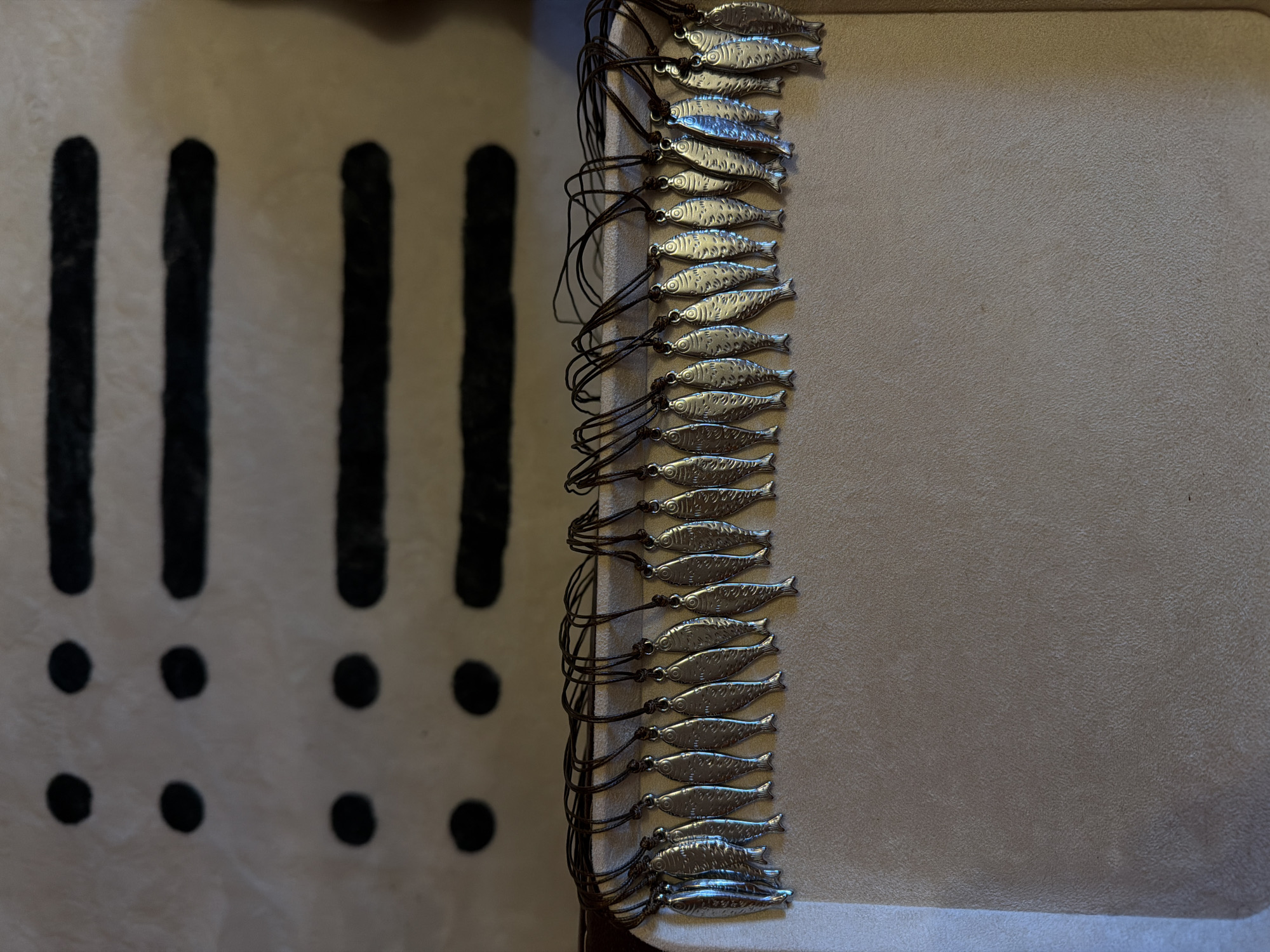 The image displays a collection of small, silver fish charms, each strung on dark cord. They are arranged in a vertical line down the right side of the frame, against a textured, light-colored surface. The charms are detailed, with visible scales and fins. To the left of the charms, on a lighter, patterned background, are four vertical black lines, each accompanied by two black dots below it, arranged in two columns. The scene is lit from the left, casting soft shadows. The overall impression is of a still-life arrangement, possibly a display of jewelry or craft items. There is no text visible in the image. The location context is Khimki, Russia.