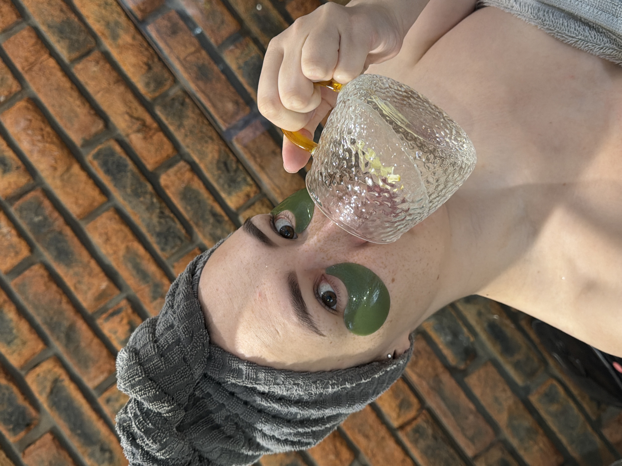 A woman lies on a brick surface, wearing a gray towel wrapped around her head and green eye patches. She is holding a clear glass cup with a yellow handle near her face. The location is potentially in the city of Chaikovsky, Russia.
