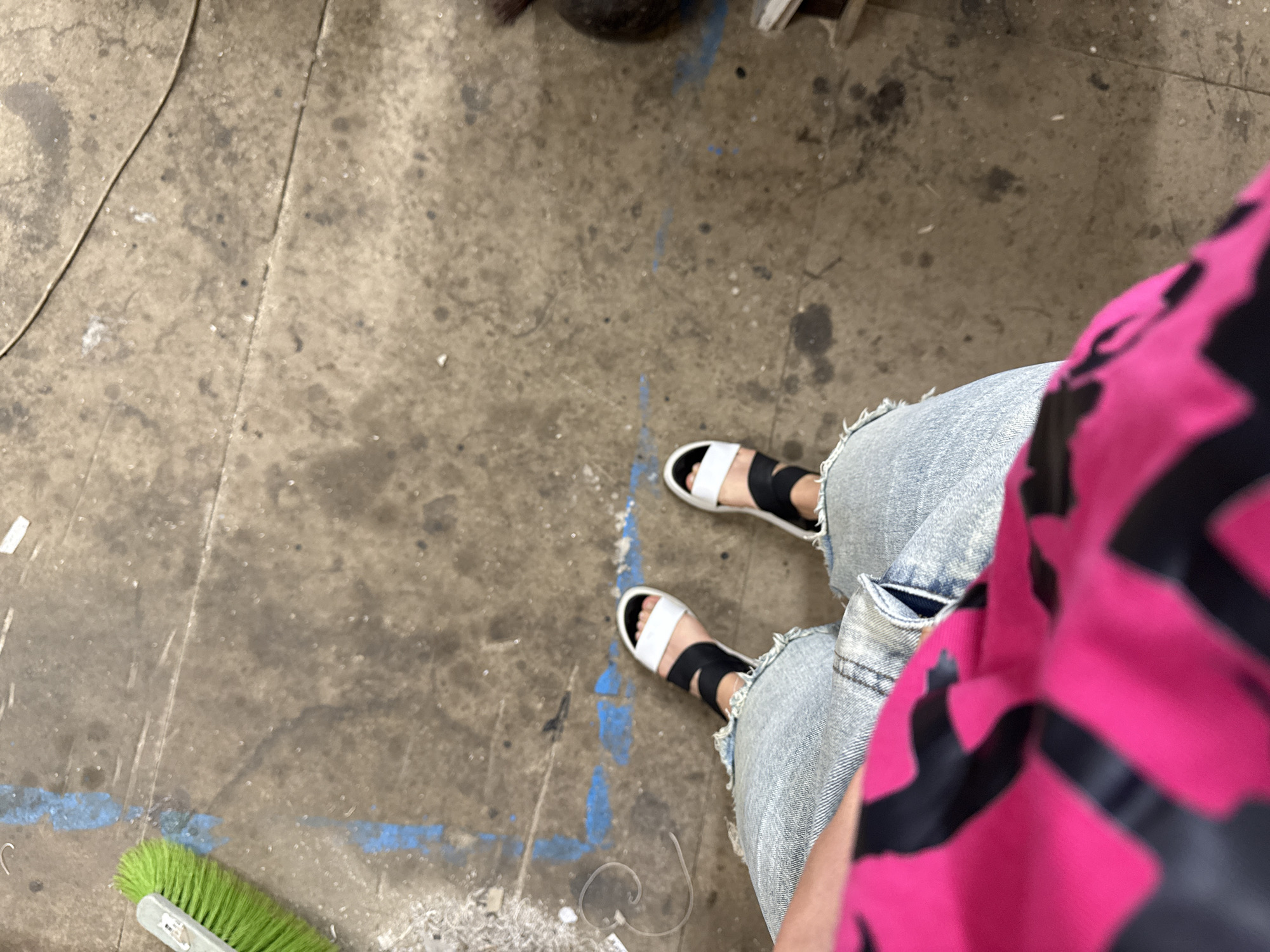 An overhead shot shows a person's legs and feet standing on a concrete floor marked with blue paint and dirt. The person is wearing light-wash ripped jeans and white and black slide sandals. Their left leg is partially covered by a bright pink and black patterned garment. A green-bristled brush lies on the floor to the left of the frame.