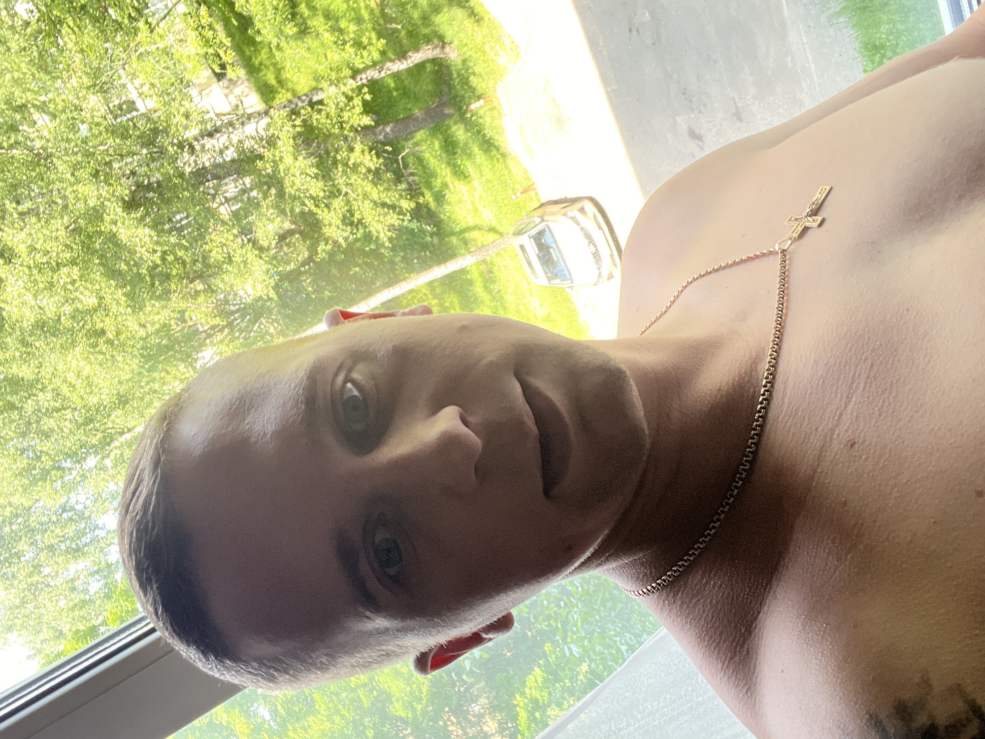 In a selfie taken from a slightly upward angle, a man with short hair and blue eyes is prominently featured. He is bare-chested, wearing a gold chain with a cross pendant. The background shows a window with white curtains on the left, and through the window, lush green trees and a white car parked on a street are visible. The overall scene suggests an indoor setting with an outdoor view, likely during daylight, with the hint of a city environment due to the parked car and implied buildings behind the trees, possibly in Minsk, Belarus.