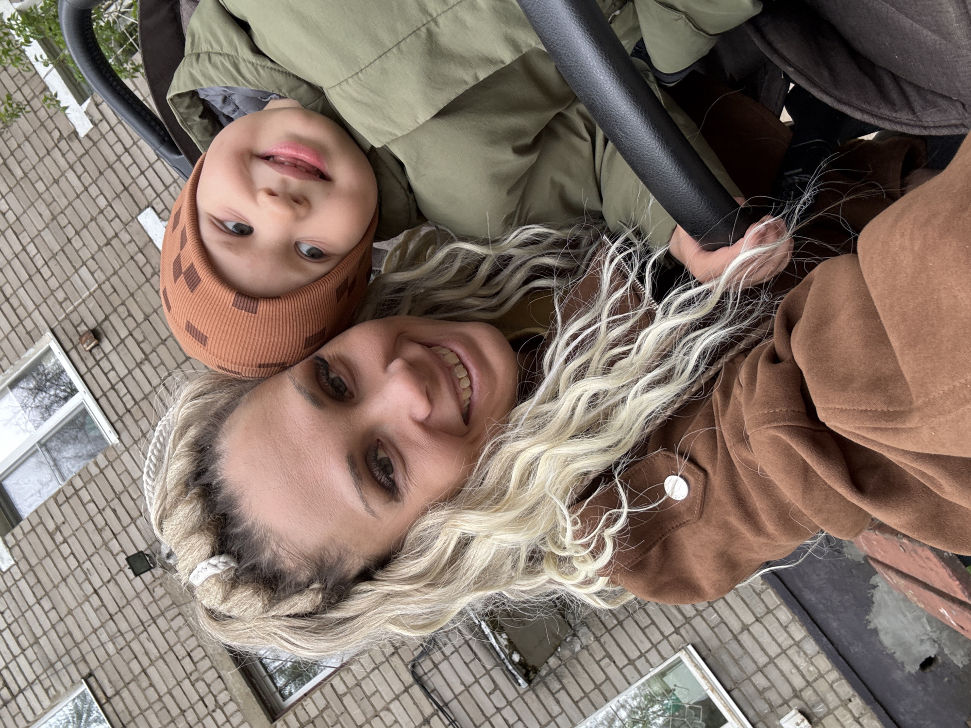 A selfie taken in the city of Rusanovo, Russia, shows a woman and a young child. The child, wearing a brown patterned hat and a green jacket, is positioned slightly above the woman. The woman is blonde with her hair in braids, wearing a brown suede jacket. The photograph is taken from a low angle, with a stroller handlebar visible in the frame. A brick building with windows forms the backdrop.
