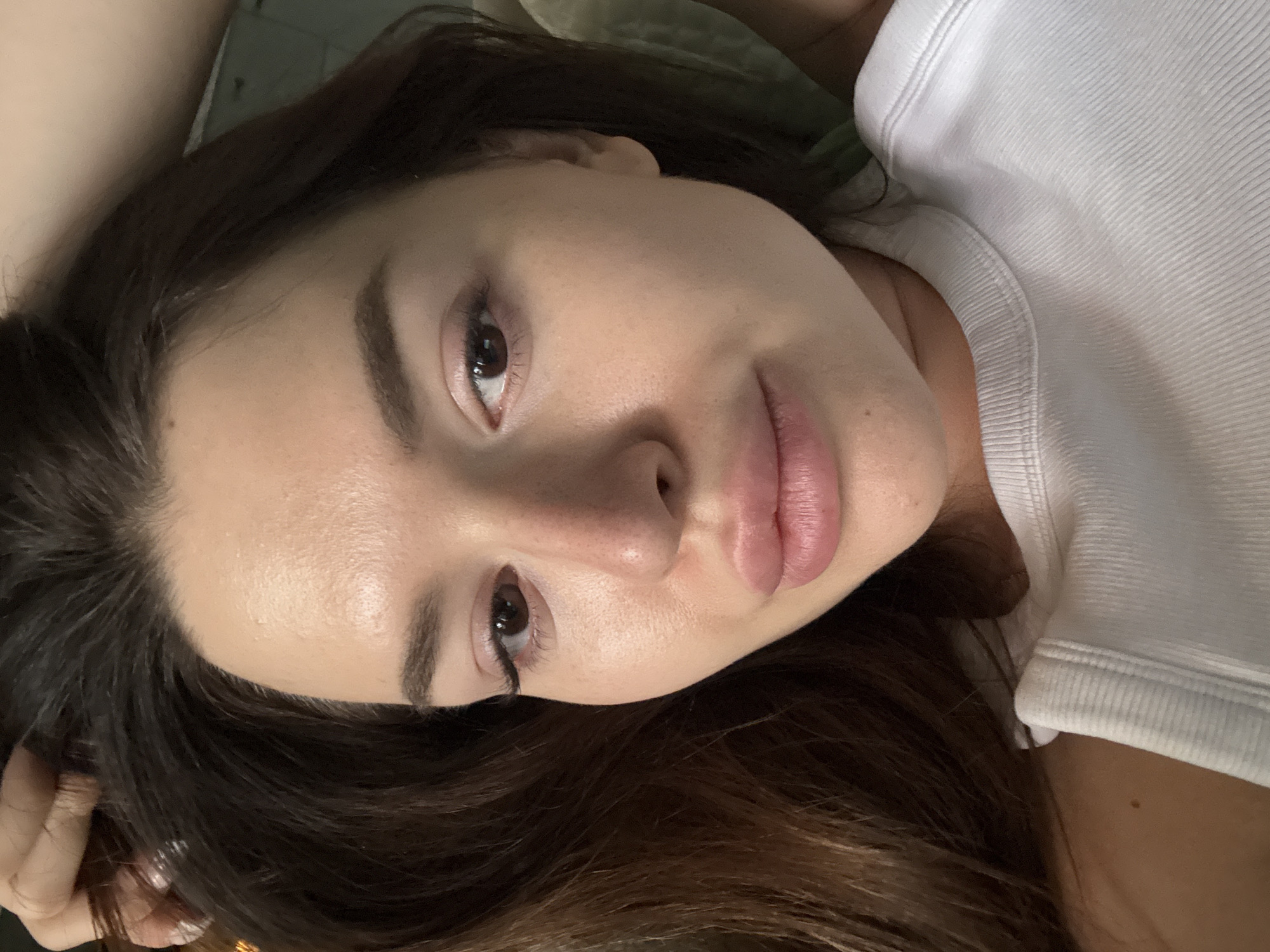 This is a close-up, overhead, selfie-style photograph of a young adult female lying down, likely on a bed, with her dark brown hair spread around her head. She is wearing a white ribbed tank top. Her head is tilted slightly to her left, and her right hand is visible at the top left of the frame, pulling some of her hair back. The lighting is soft and diffused, creating a natural glow on her skin. Her face is the primary focus, with her eyes open and looking upwards. She has noticeable lip filler. The background is out of focus and appears to be a light-colored sheet or blanket. The overall impression is one of a casual, intimate moment, captured in a domestic setting. There are no indicators of the specific location being Prague, Czechia.