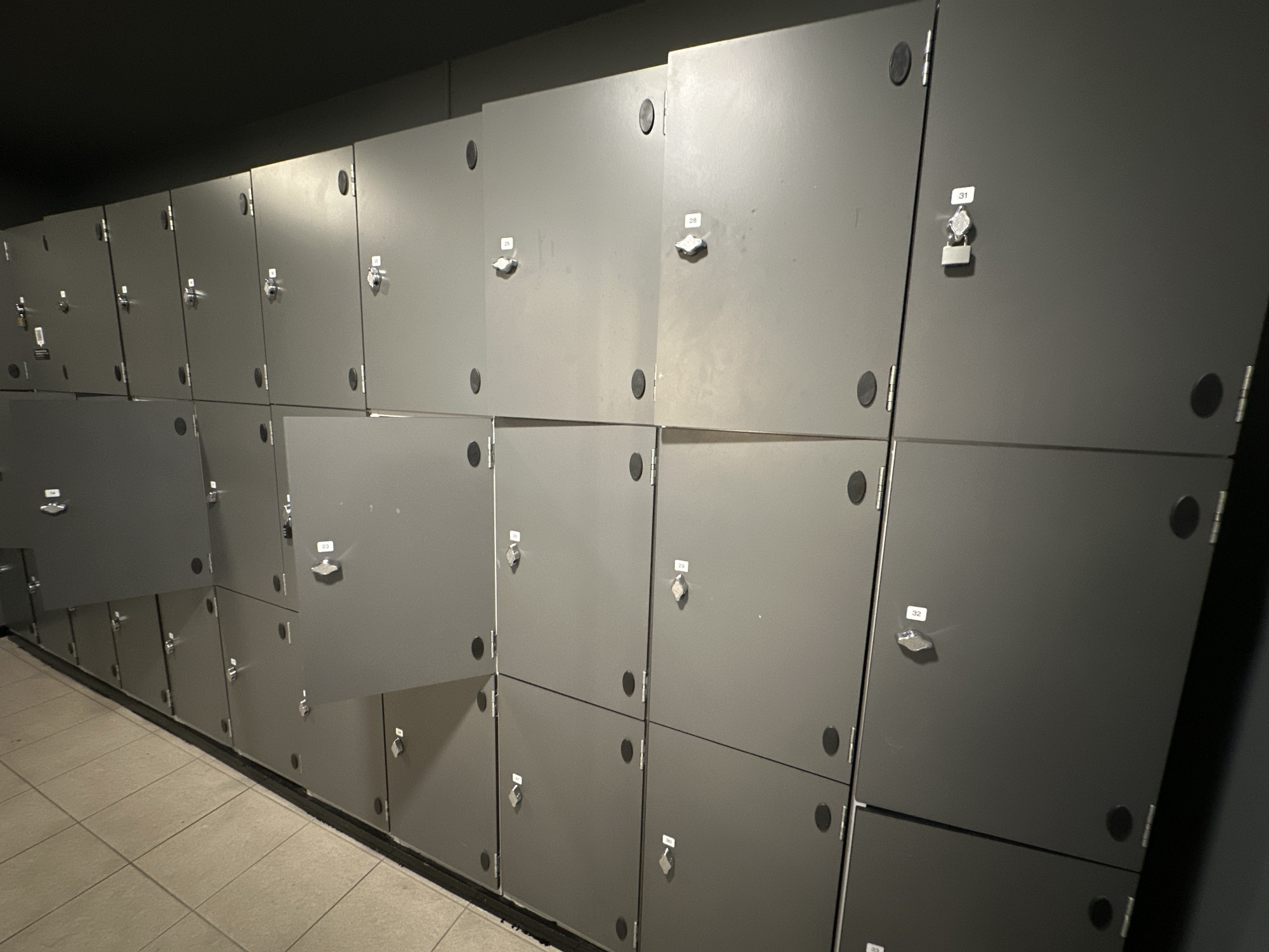 The image shows a row of gray metal lockers. Each locker has a number on it and a lock. Some of the lockers are open, while others are closed. The lockers are arranged in two rows, one above the other. The floor in front of the lockers is tiled with light-colored square tiles. The lockers appear to be in a public place, such as a gym, pool, or changing room. Without further information or context, it is impossible to determine the specific location in Carlow, Ireland.