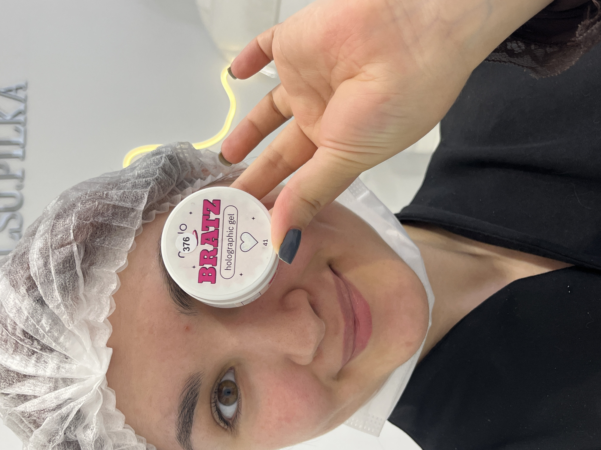 A young adult female is depicted in a close-up, upper-body shot, smiling slightly and facing the camera. She wears a white disposable hairnet and a white disposable face mask positioned below her chin. Her visible attire consists of a black garment, likely a smock or robe, with dark lace detailing on her right sleeve. Her left eye is brown and visible; her right eye is obscured by a small, white, circular container held in her right hand. The container's label features prominent pink text 'BRATZ holographic gel,' along with the number '376' and a heart icon above '41.' Her fingernails are painted dark grey. The background is a white wall featuring raised silver lettering that partially spells 'L.SU.PILKA' in the upper left, and a section of a glowing yellow neon light on the right. This scene is situated within the L.SU.PILKA nail salon in Neftekamsk, Russia.