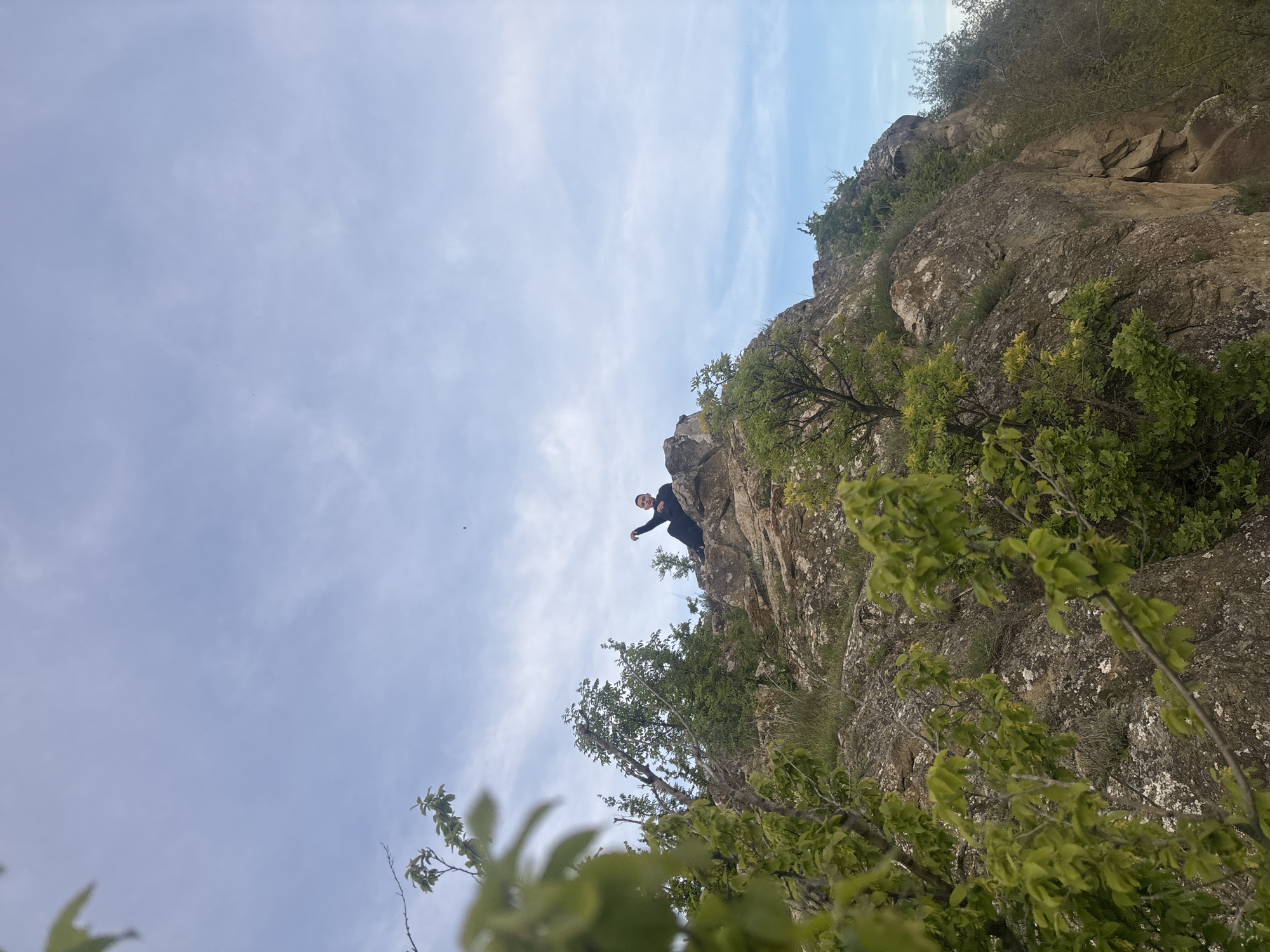 A low-angle shot reveals a person perched on a rocky cliff in Goritsa, Bulgaria. The sky is partially covered with scattered white clouds, with the rocky cliff dominating the right side of the frame. The person is wearing dark clothing and appears to be reaching out or gesturing with their arm. Green foliage and trees grow on the cliff face, partially obscuring the rocks. The composition suggests the photographer is positioned below, looking upward at the person on the cliff.