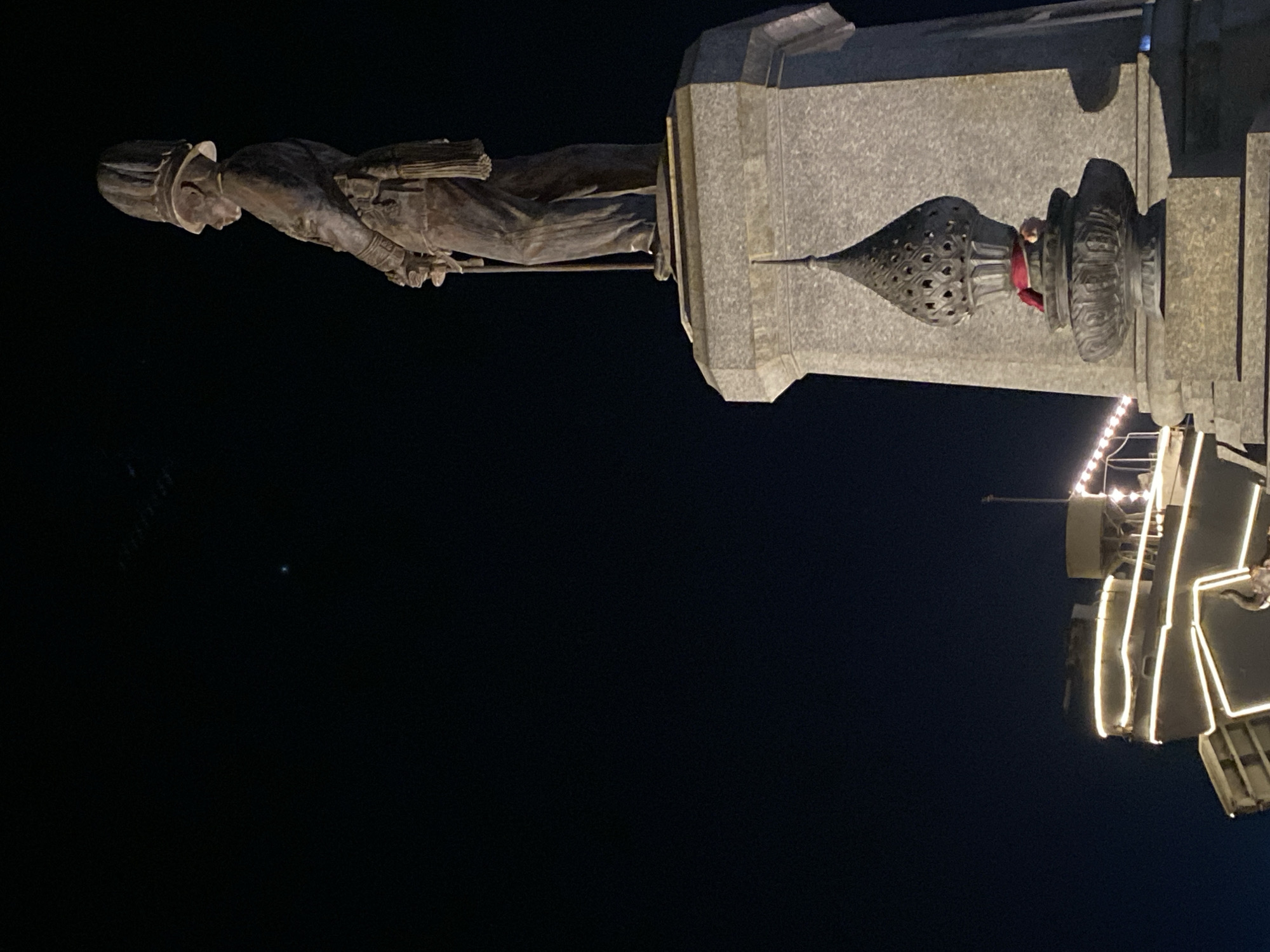 At night, a low-angle, medium shot captures a statue mounted on a stone pedestal. The statue, rendered in grayscale, depicts a man wearing a hat and holding a staff, possibly a depiction of a historical figure. The base of the statue features an ornate, rounded pinnacle. To the right of the statue, a series of glowing, linear light fixtures is visible. The dominant background is a vast expanse of darkness, indicative of the night setting. Based on the visual cues, the statue is located in Baan Don Sai, Thailand.