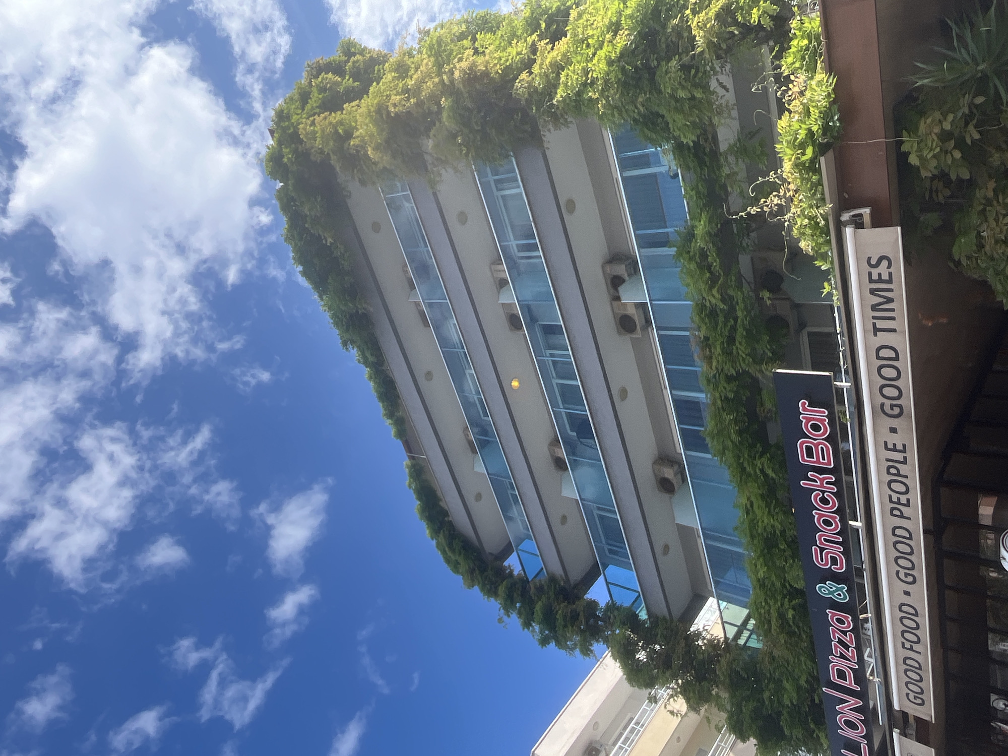 The image is an upward-angled view in Alanya, Turkey, featuring a modern building covered in greenery, juxtaposed against a bright blue sky dotted with fluffy white clouds. The building has multiple floors, each with large blue-tinted windows, and air conditioning units visible on the facade. The greenery, likely vines or climbing plants, drapes from the top and sides of the building. A sign for "LION Pizza & Snack Bar" is partially visible on the right side of the frame, advertising "GOOD FOOD - GOOD PEOPLE - GOOD TIMES".
