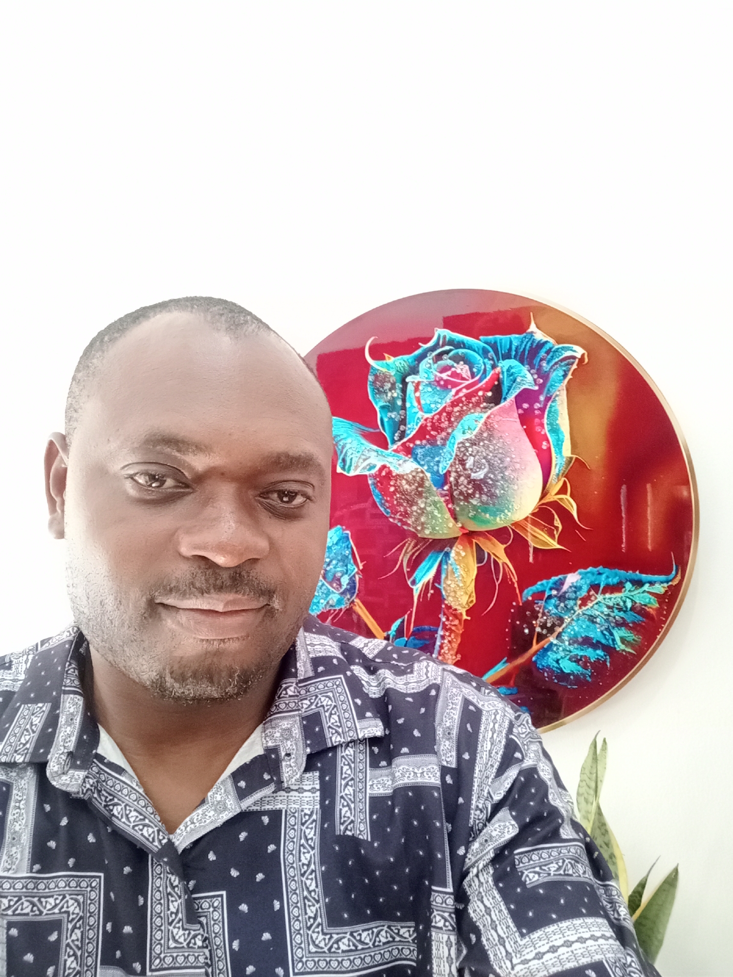 A man is photographed indoors in Gwarinpa, Nigeria. He is an African male with a bald head, light beard and a patterned shirt. Behind him is a round artwork depicting a multi-colored rose with red, blue, and yellow tones. A plant with green leaves is also visible behind the man. The background is a white wall.
