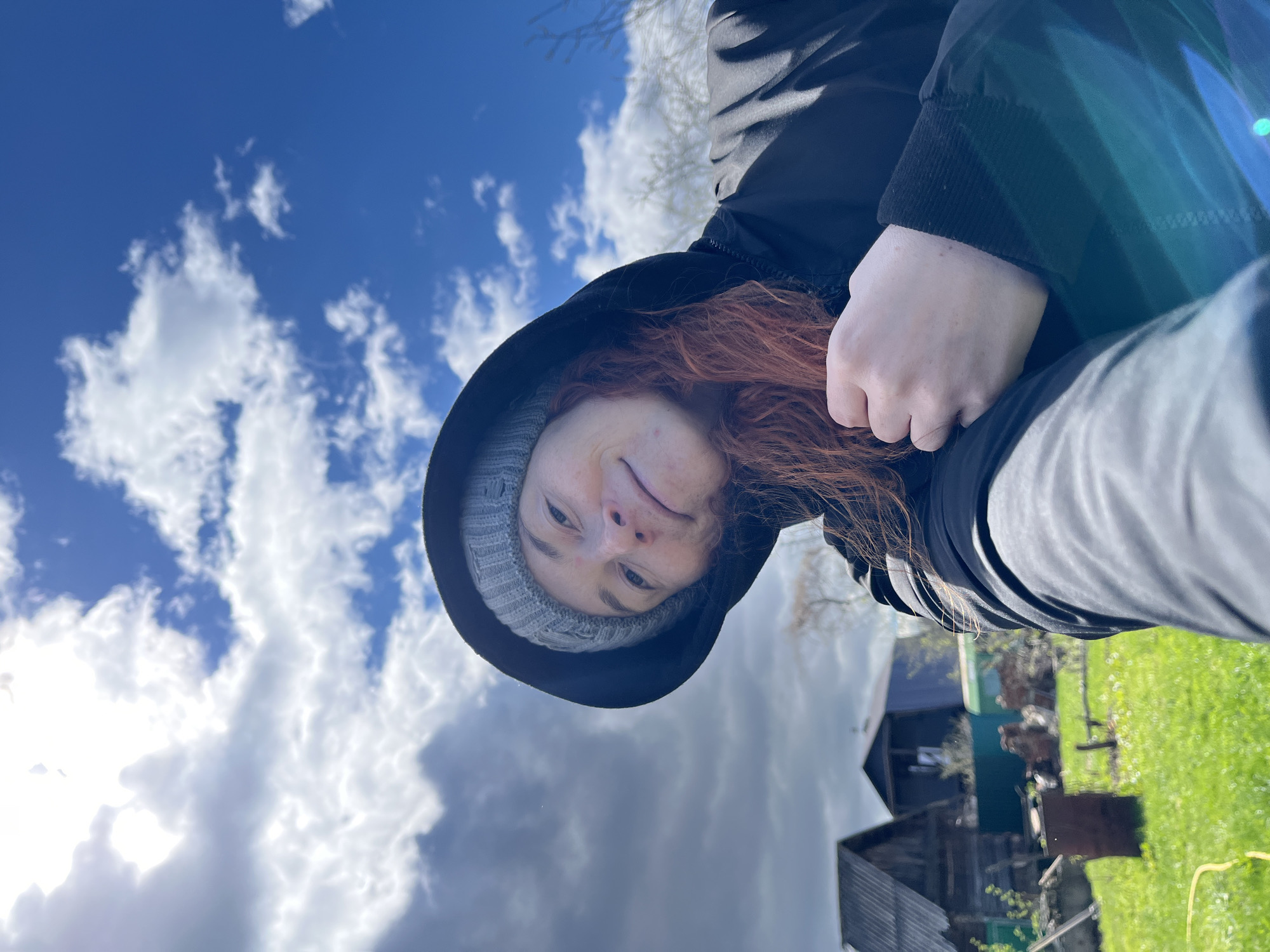 A woman with red hair, wearing a gray knit cap and a black hooded jacket, is photographed outdoors. The background features a blue sky with white clouds. In the lower portion of the image, there is a building and green grass. The image seems to be taken with a slight upward angle and possibly in the city of Chaikovsky, Russia.
