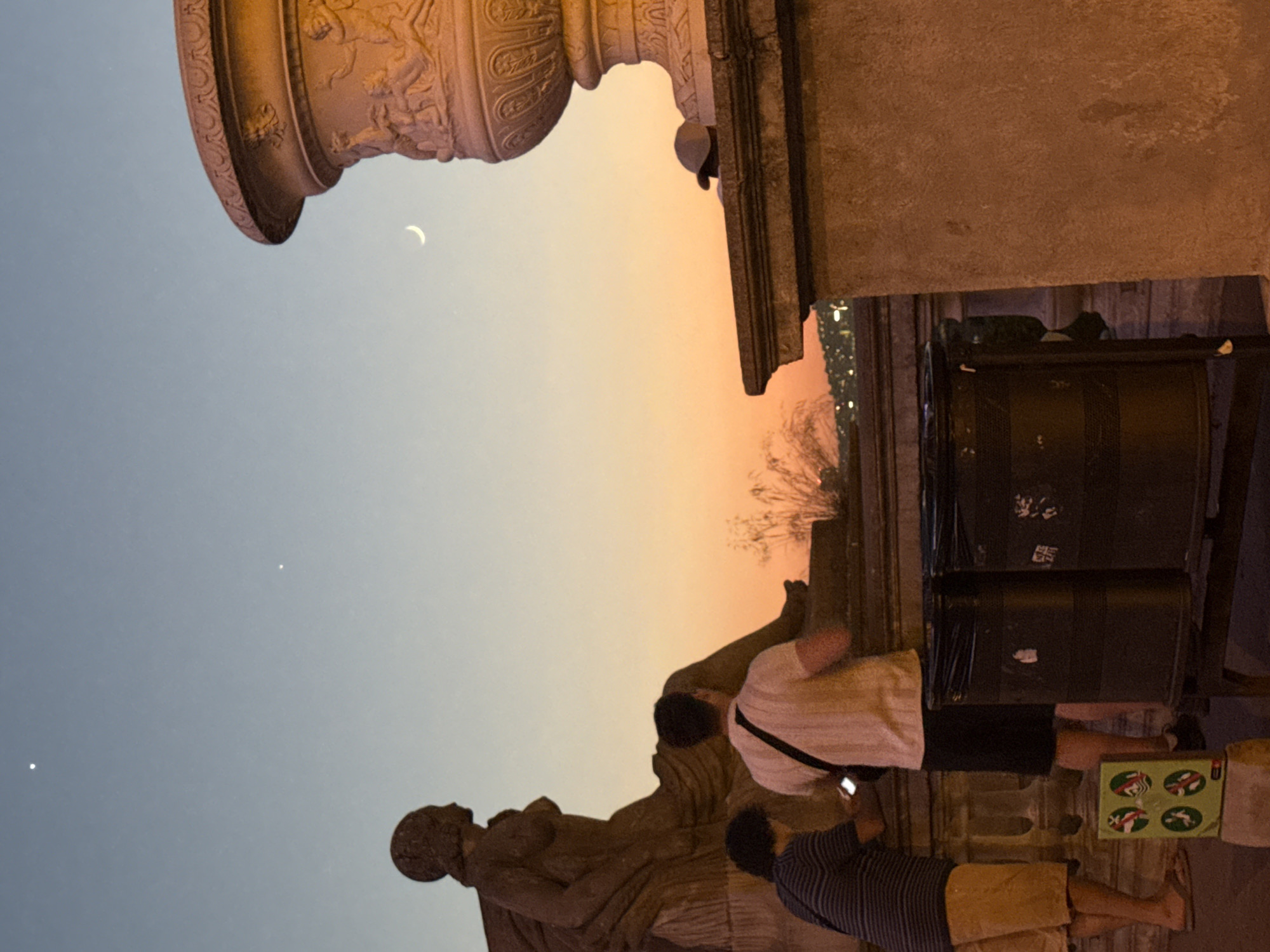 The image depicts a twilight scene at Montjuïc Castle in Barcelona, Spain. Two individuals are visible in the foreground, one wearing a striped t-shirt and khaki shorts, the other in a white t-shirt and dark shorts. The individual in the striped t-shirt is holding a smartphone. They are standing near two dark trash receptacles. Behind them, a large stone statue of a seated female figure is prominent. To the right, a large decorative stone urn is partially visible. The sky displays a gradient of colors from pale blue at the top to orange and yellow near the horizon, indicating either sunrise or sunset. Two bright celestial bodies, likely stars or planets, are visible in the upper left portion of the sky, and a crescent moon is visible in the upper right. In the background, a cityscape with lights is visible in the distance.