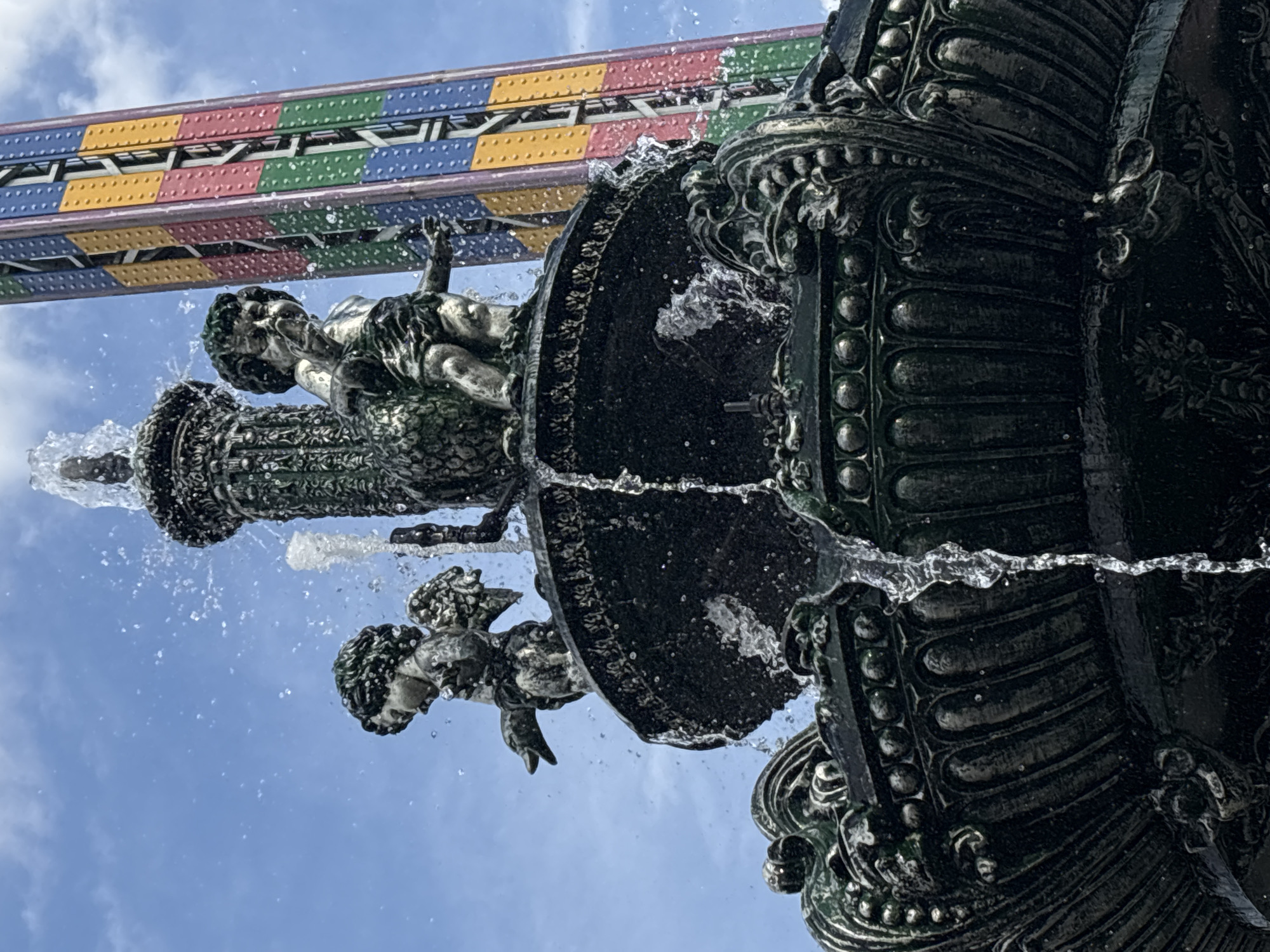 This is a close-up, low-angle shot of a decorative fountain in Yerevan, Armenia. The fountain features dark-colored, ornate sculptures of cherubs with wings. Water is actively spraying and cascading from the fountain, creating a dynamic and wet effect. In the background, a tall, brightly colored structure, possibly a ride from an amusement park, with sections of blue, green, yellow, and pink, is visible against a cloudy blue sky. The composition focuses on the interplay of the water, the sculpted figures, and the colorful backdrop.