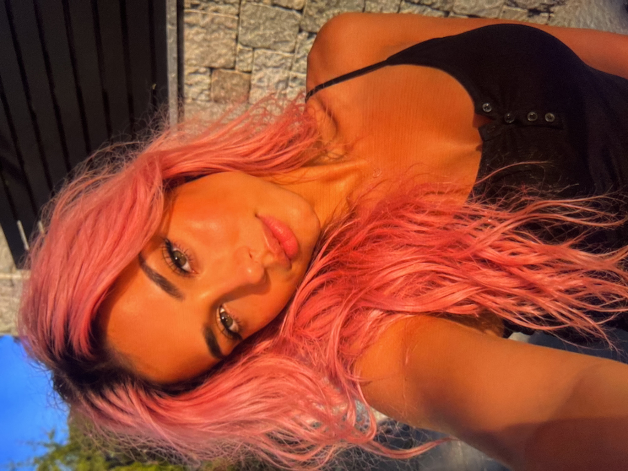 A young woman with bright pink hair is taking a selfie outdoors. She is wearing a black tank top. The background consists of a stone wall and a dark structure, possibly a building, against a bright blue sky. The lighting suggests it is either sunrise or sunset, casting a warm, orange glow on her skin and hair. Her expression is relaxed and she is looking directly at the camera.