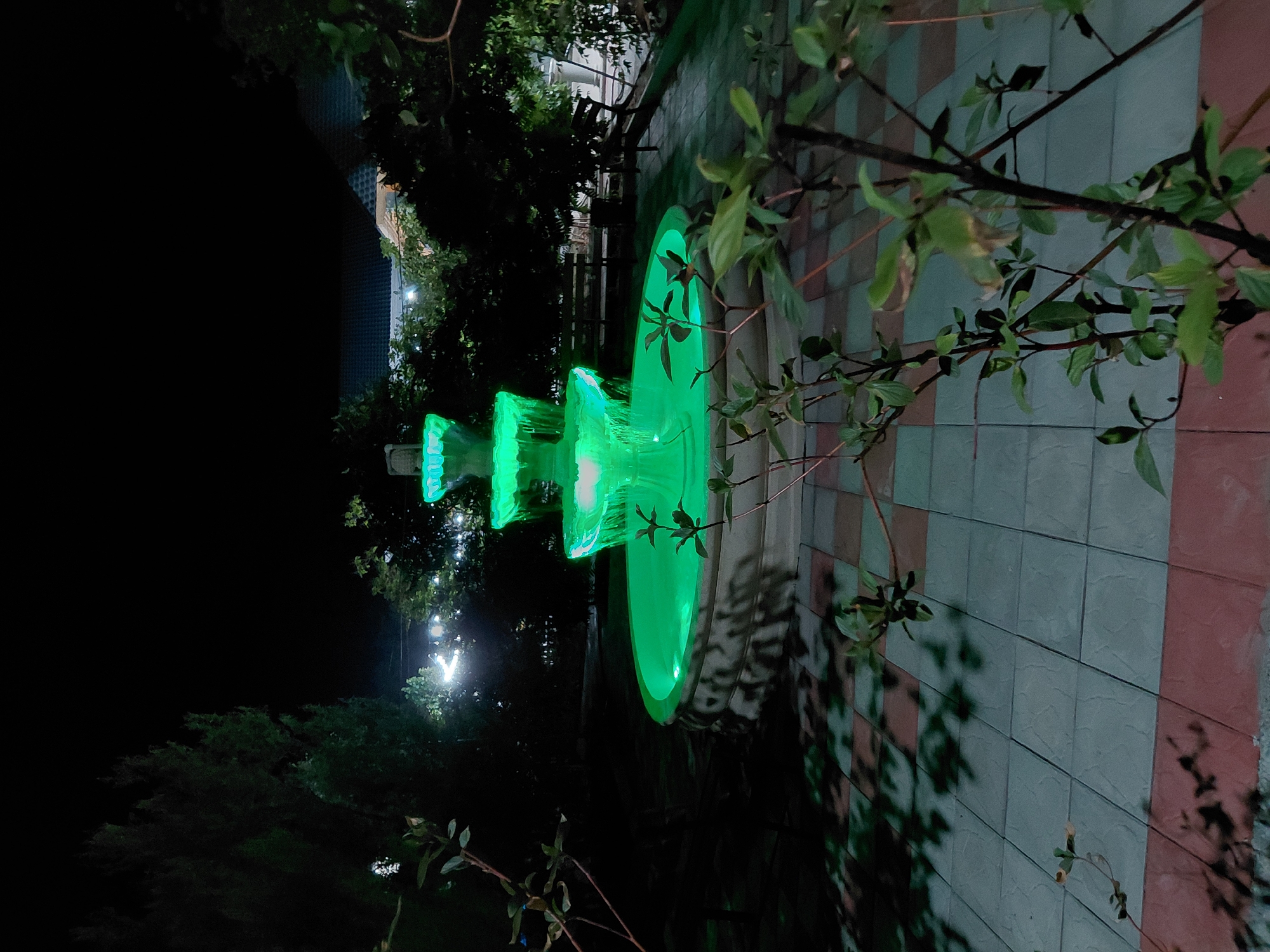 This image depicts a night scene in Akschi, Kazakhstan. The primary subject is a modern, multi-tiered fountain illuminated with vibrant green LED lights. The fountain is set against a backdrop of lush greenery and a tiled wall. The time of day is clearly nighttime, as indicated by the darkness and the prominent artificial lighting of the fountain.
The fountain's tiered design features cascading water illuminated from within, creating a glowing effect. The largest tier is at the bottom, with progressively smaller tiers above it, creating a dynamic visual. The surrounding area appears to be an outdoor space, possibly a garden or courtyard, with foliage from trees and plants visible in the foreground and background. The tiled wall behind the fountain has a pattern of light and dark grey squares interspersed with pinkish-red rectangles.
Subtle details like the shadows cast by the plants on the wall and the soft glow of distant lights in the background suggest a calm and perhaps serene atmosphere. There are no people visible in the image, focusing all attention on the illuminated fountain and its environment. The weather appears to be clear, with no rain or strong winds suggested. The image is oriented vertically, with the fountain appearing in the lower right quadrant when viewed in a standard landscape orientation. No visible text is present in the image.