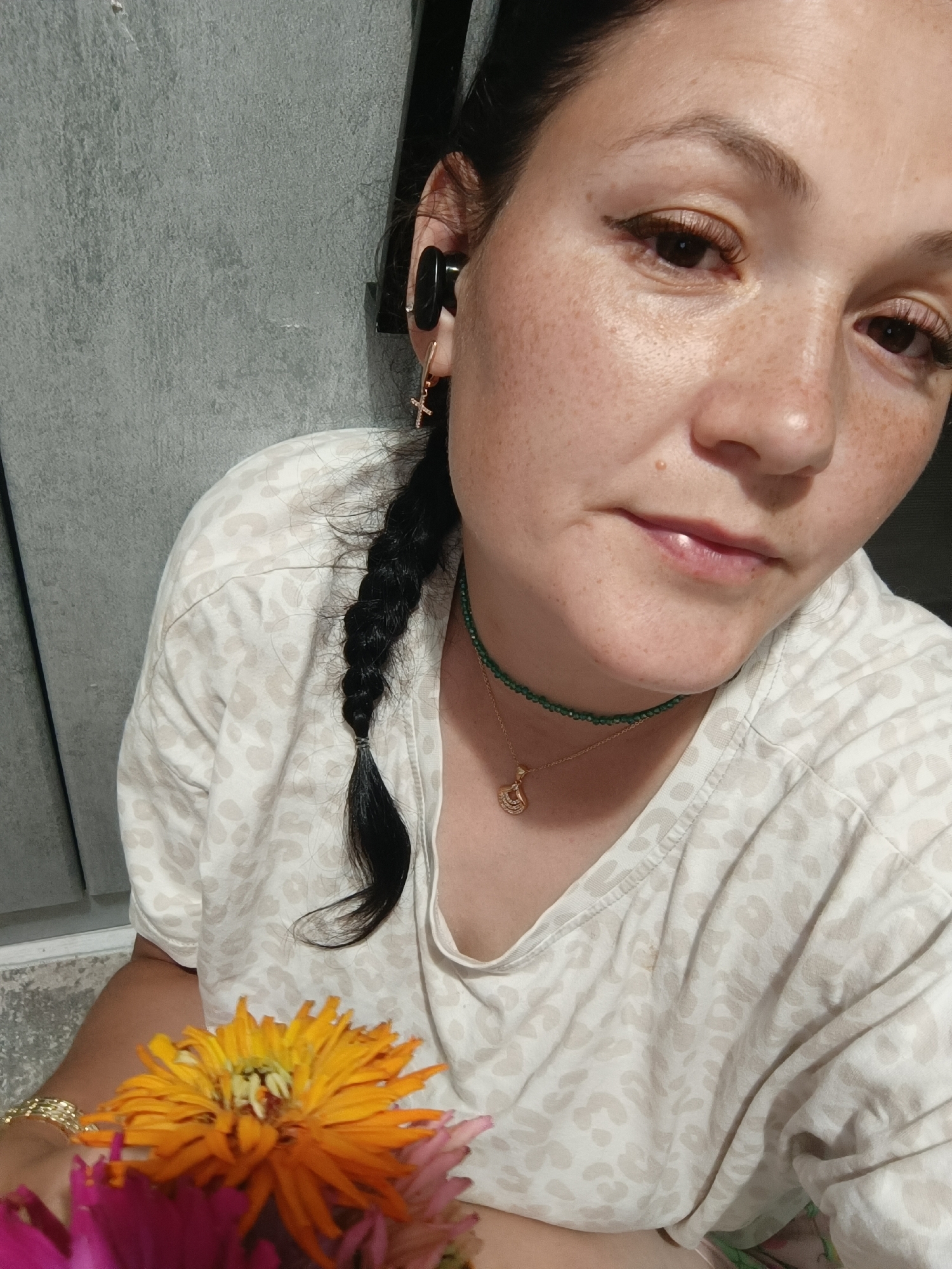 A close-up selfie shows a woman with dark hair in a braid, wearing a light-colored shirt with a leopard-print pattern. She is wearing a green beaded necklace with a gold pendant and small hoop earrings, one of which has a cross charm. She has dark eyeliner and light freckles across her face. She is holding a bouquet of flowers, including an orange chrysanthemum and pink dahlias. The background appears to be a grey, textured wall, possibly an exterior wall of a building. The image is cropped tightly, focusing on the woman's face and upper body. The lighting suggests it is daytime.