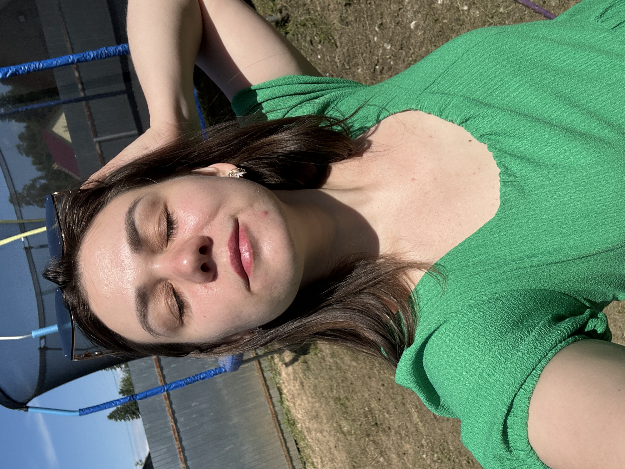 In a candid outdoor shot, a young woman with brown hair closed eyes and her hand on her head, enjoys the sun. She is wearing a green dress and sunglasses. The background shows a trampoline and a fence, hinting at a backyard setting, possibly in the city of Chaikovsky, Russia, where a house and trees are visible in the distance. The overall ambiance is relaxed and serene.