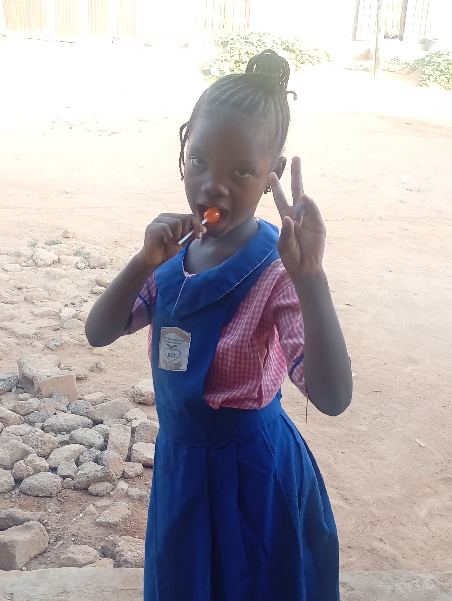 A young, dark-skinned girl stands outdoors. She wears a pink and white checkered short-sleeved shirt under a dark blue dress with a badge. The badge appears to have a logo with a heart and the letters "BET". She is eating an orange lollipop and holding up two fingers in a peace sign. Her hair is styled in braids. The background is a sandy or dirt ground, with rocks and debris scattered around. In the distance, there's a building or structure with a light-colored wall. The location is in the city of Unknown, Unknown.