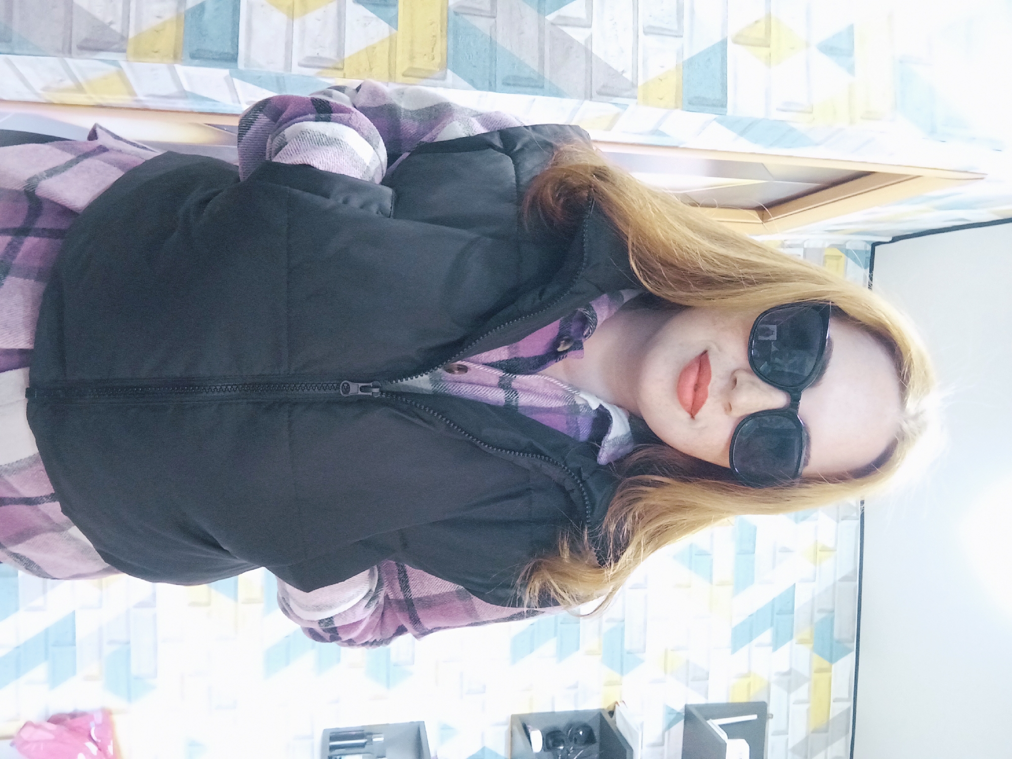 The image shows a woman wearing sunglasses, a black vest, and a purple and white plaid shirt. The location appears to be indoors, based on the wall with a geometric pattern and the presence of furniture. There is no information to suggest that the image was taken in Yurgamysh, Russia. The description of the location should remain within the image's context, not be based on assumptions.