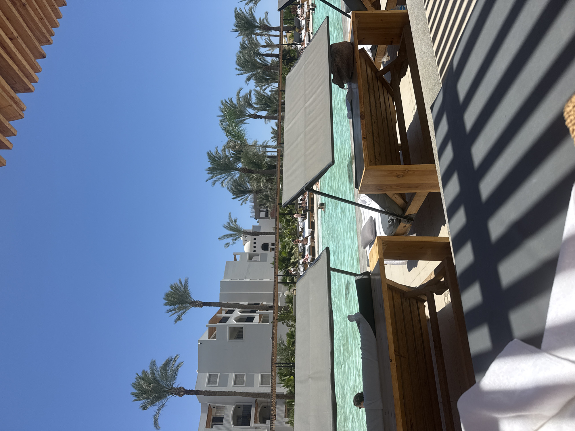 A sunny day at a resort pool in Hurghada, Egypt. The image shows lounge chairs with canopies by the pool, a building in the background with balconies and palm trees. People are visible relaxing on lounge chairs and in the pool. The sky is clear blue, and sunlight casts shadows from an overhead structure onto the foreground lounge chairs.