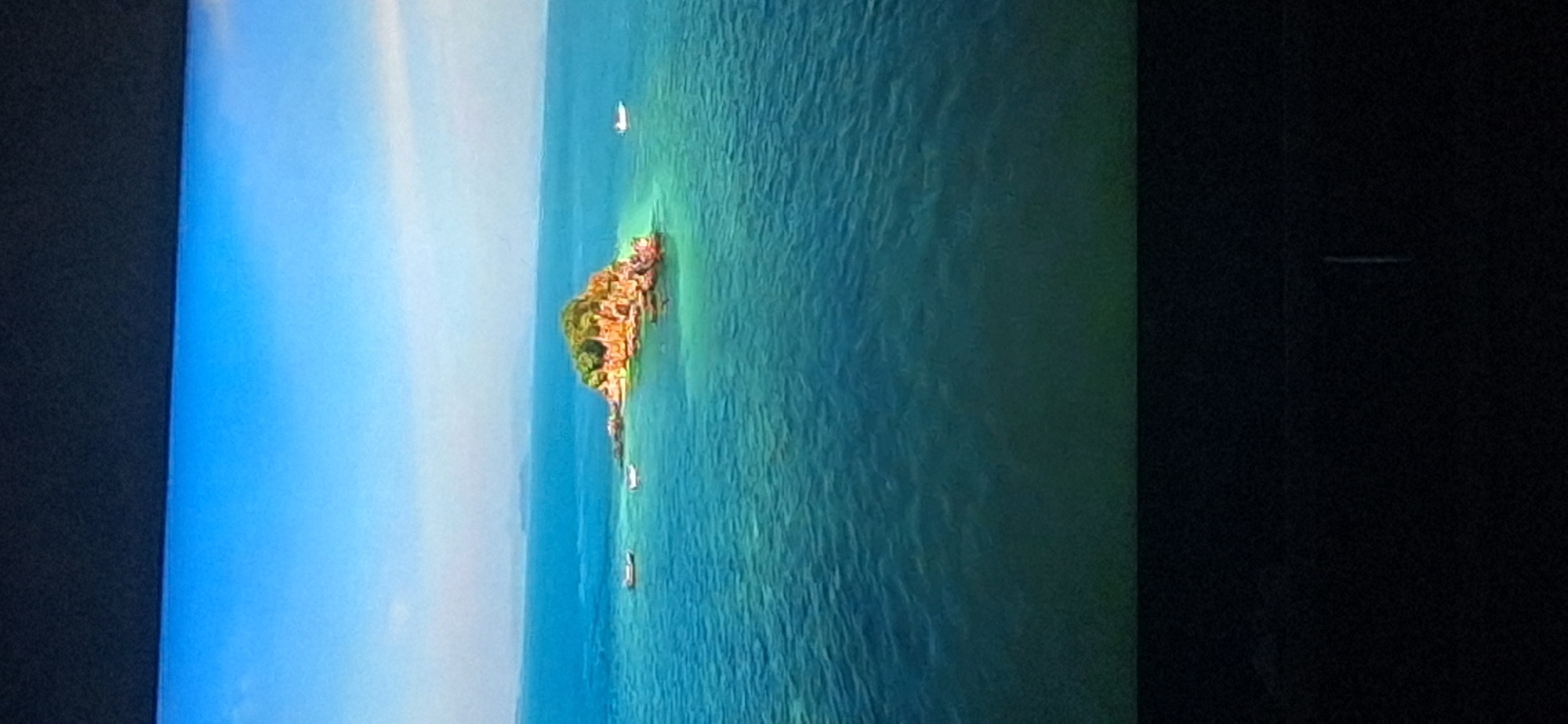 The image depicts an aerial view of a small, rocky island with lush green vegetation on top, situated in clear turquoise water. Several small boats are visible in the water surrounding the island. The sky is a vibrant blue with some hazy clouds on the horizon. The lower portion of the image shows a dark, out-of-focus area which appears to be the bottom of the screen or frame. There is no identifiable information within the image to suggest it is located in Bila Tserkva, Ukraine.