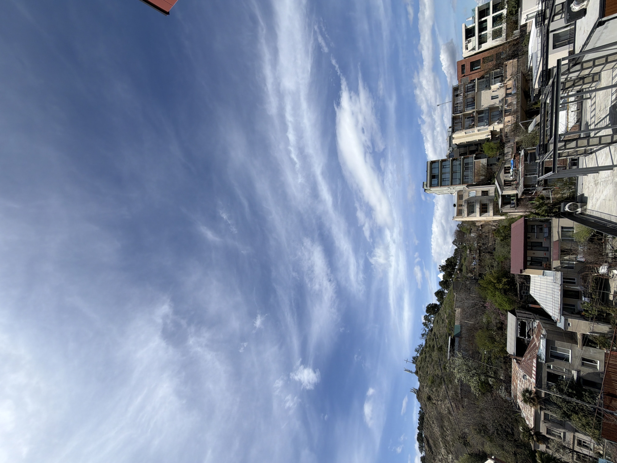 The image captures a daytime scene in Tbilisi, Georgia, featuring a view of a hillside cityscape under a partly cloudy sky. The sky is predominantly blue with streaks and patches of white clouds running diagonally across the frame. The urban landscape is characterized by a mix of residential buildings, including multi-story apartments and smaller houses, some with red or gray roofs. The buildings are arranged along a hillside, interspersed with green vegetation and trees. The architectural styles appear varied, reflecting a mix of old and modern constructions. A portion of a metal structure is visible in the foreground, suggesting a balcony or terrace. The light suggests a sunny day, casting shadows that provide depth and texture to the environment.