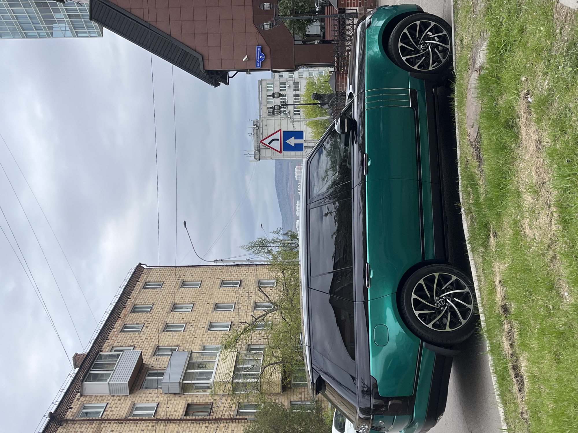 The image depicts a street scene in Krasnoyarsk, Russia. A green Range Rover is parked on the side of the road. In the background are apartment buildings, an overhead street sign indicating a traffic turn, and a street sign that reads "ул. Ломоносова, 1" (Lomonosova Street, 1). The sky is overcast. Grass is visible along the edge of the sidewalk.